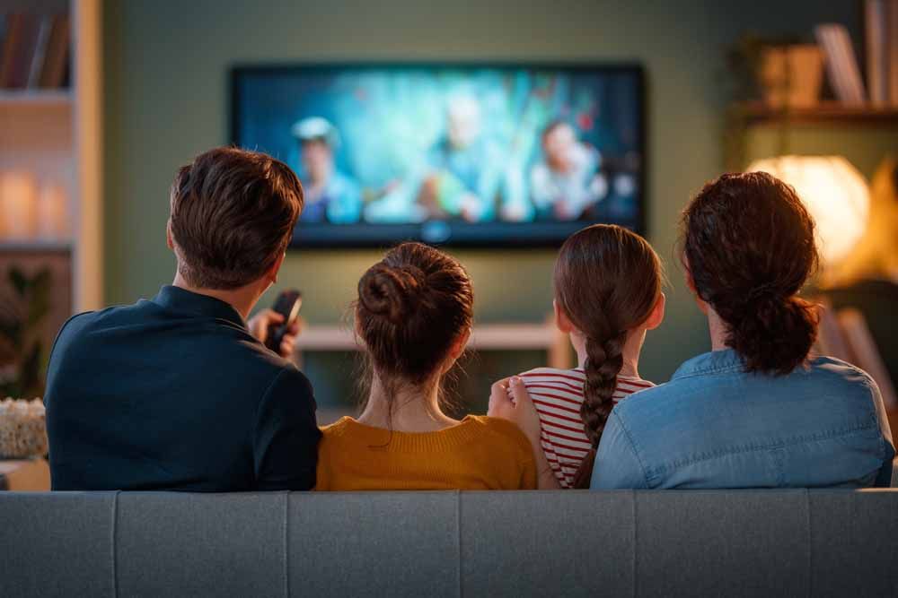Family watching TV on a couch: blue, yellow, and pink shirts; green wall, wooden shelf.
