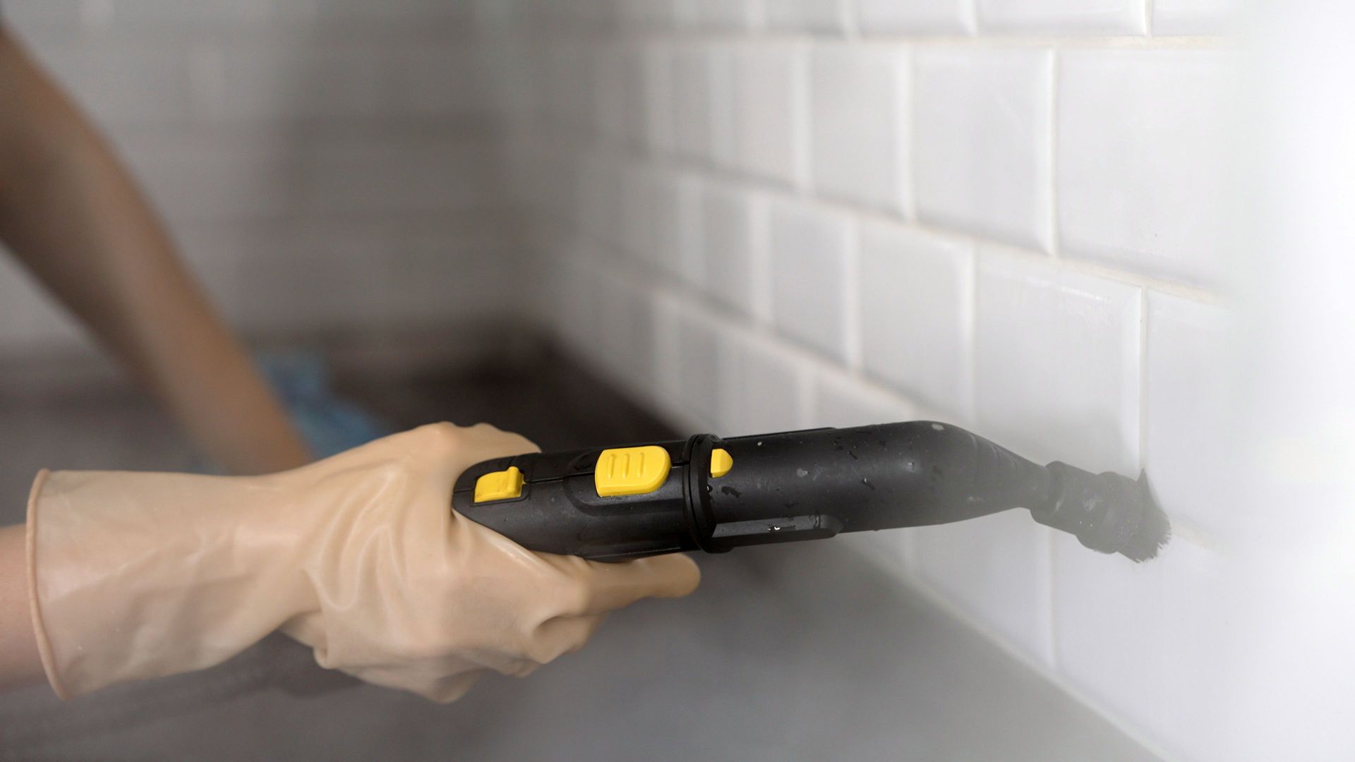 Person wearing gloves uses a steam cleaner on white tile wall and countertop.