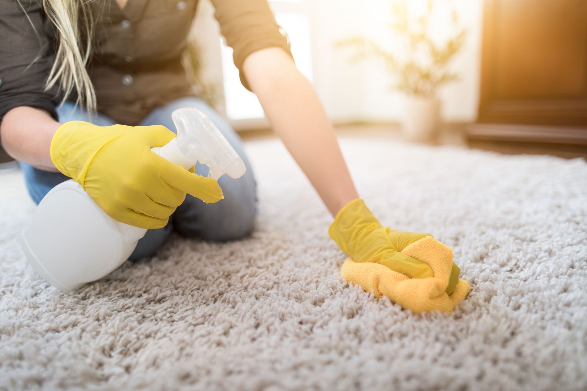 Person cleaning a cream-colored rug with yellow gloves, spray bottle, and sponge. Sunlight streams through window.