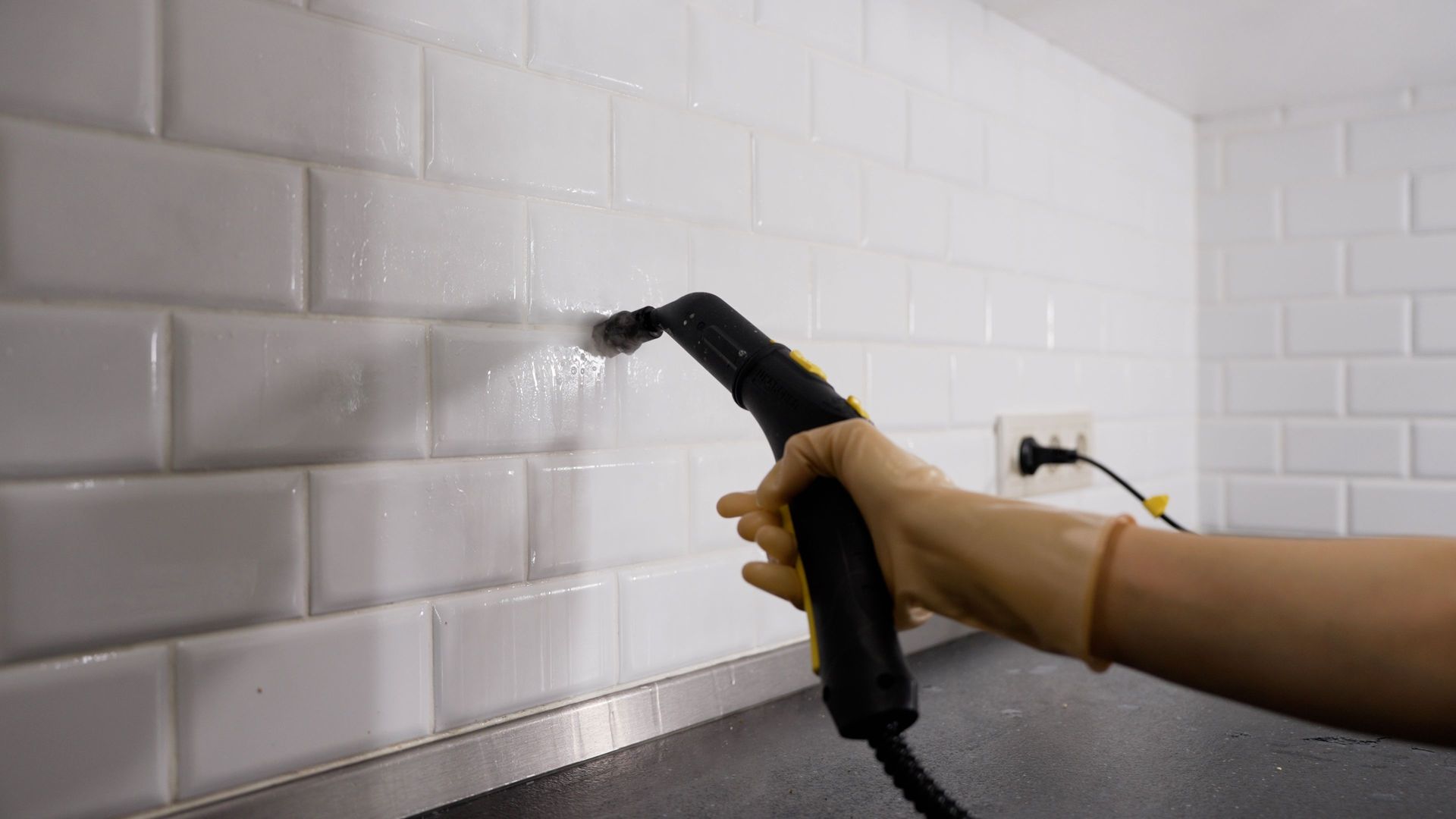 Gloved hand using a handheld steam cleaner on grout lines of a white subway tile wall