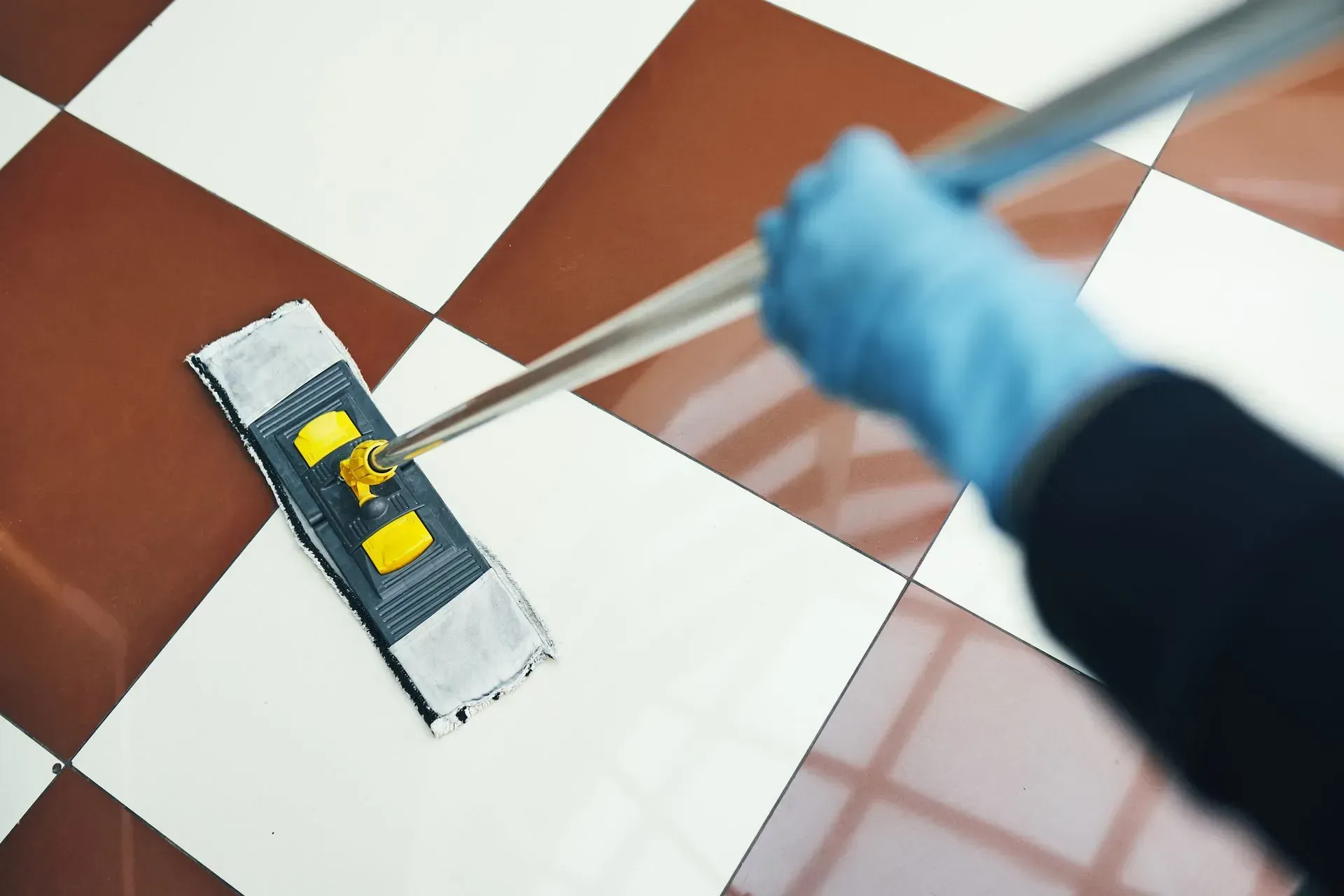 A cropped view of a person cleaning a tiled floor using a mop and wearing a blue protective glove.