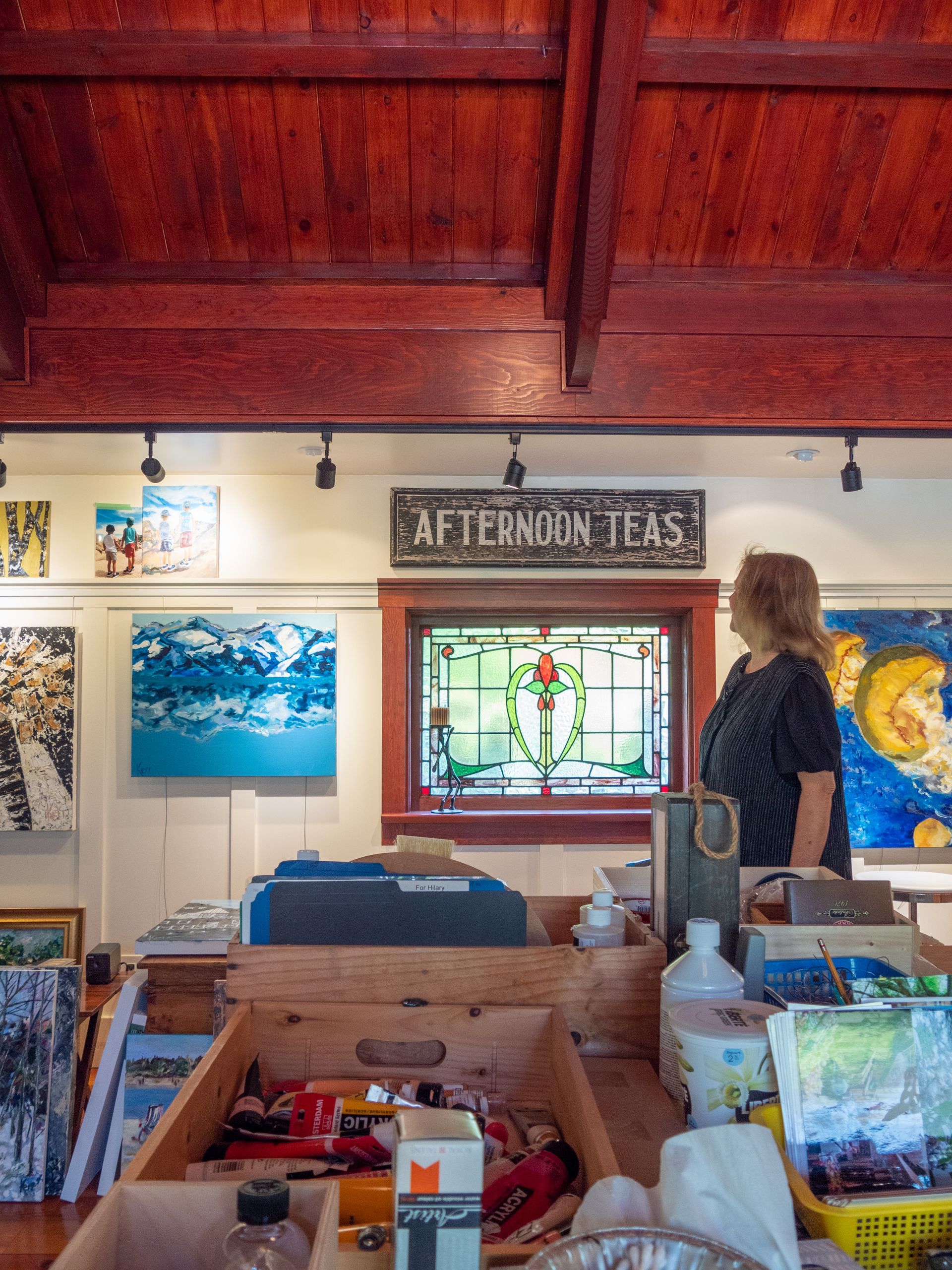 Hilary Kerr inside her studio, looking at the wall with art hanging around her and a artists table with various art tools and paint. 