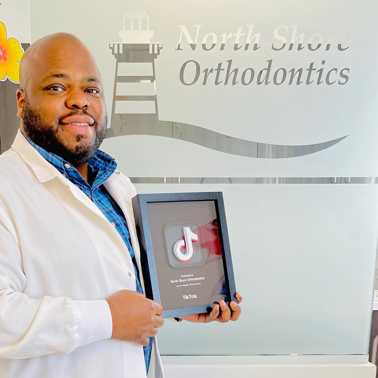 A man in a white coat is holding a framed picture in front of a north shore orthodontics sign.