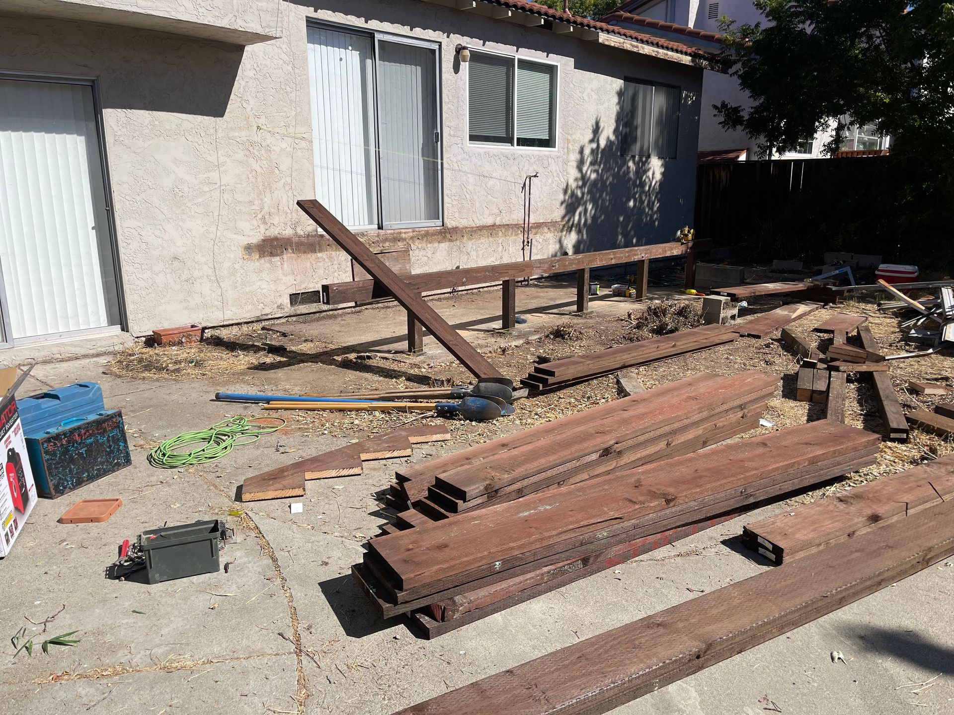 A house exterior with an unfinished wooden deck frame and piles of wooden lumber scattered on a concrete patio.