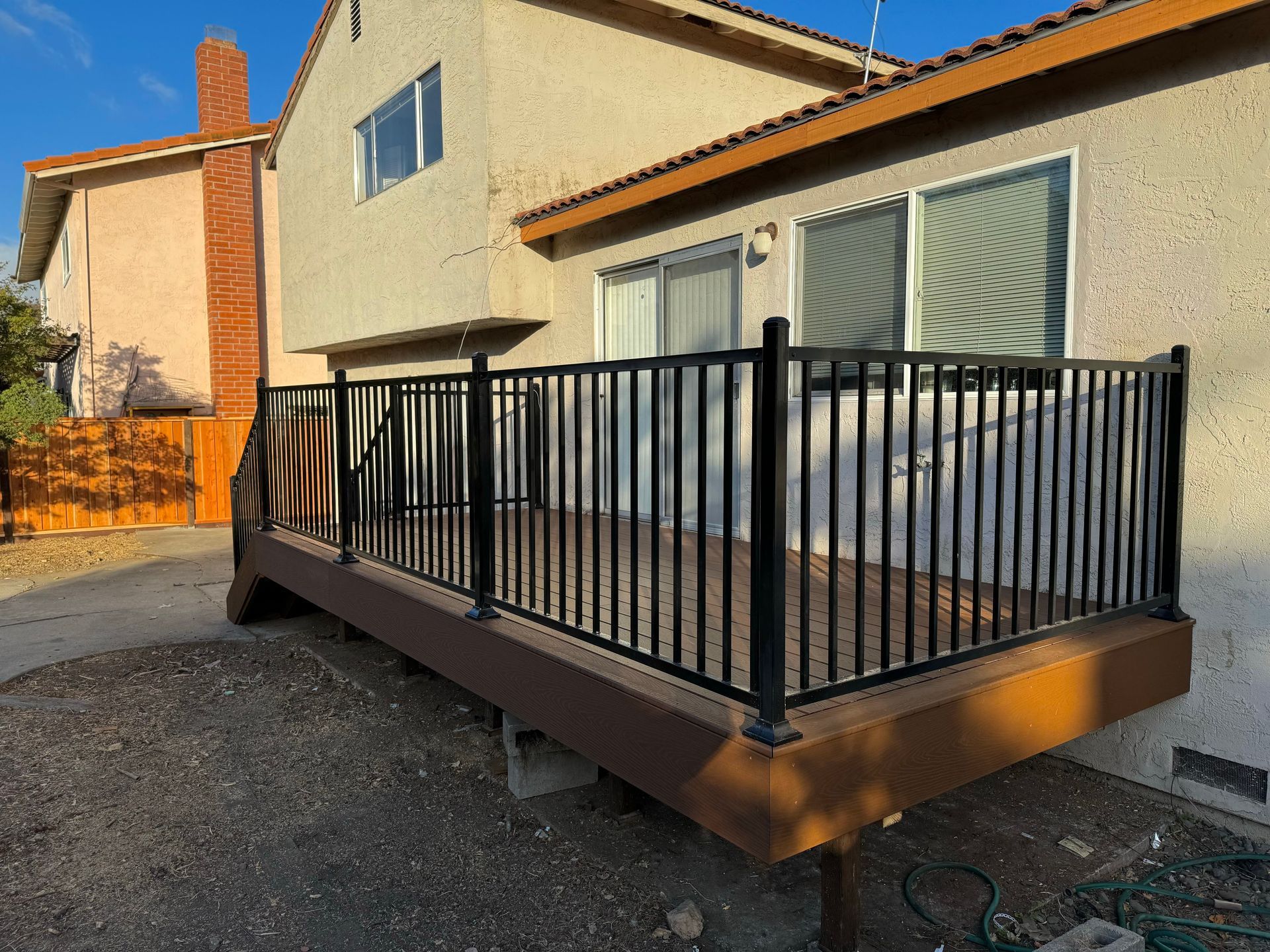 A brown deck with black metal railings attached to the side of a beige two-story house under a clear blue sky.