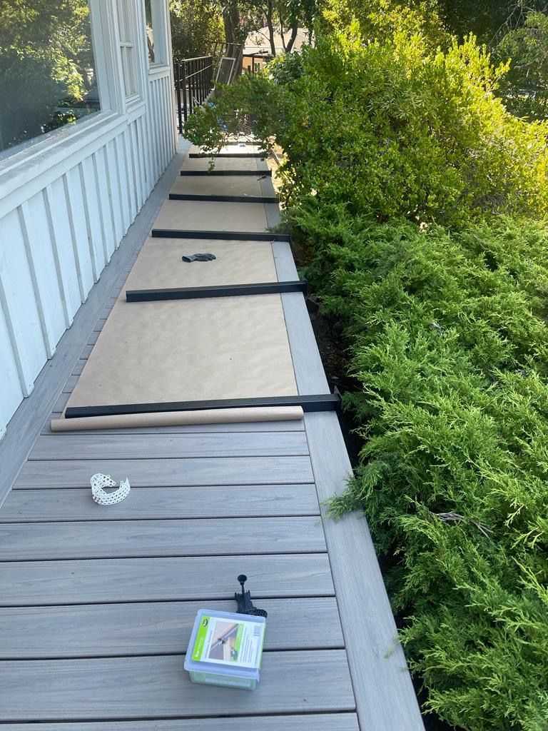 A strip of brown protective paper is rolled out along a grey composite deck next to a row of shrubs and house wall.