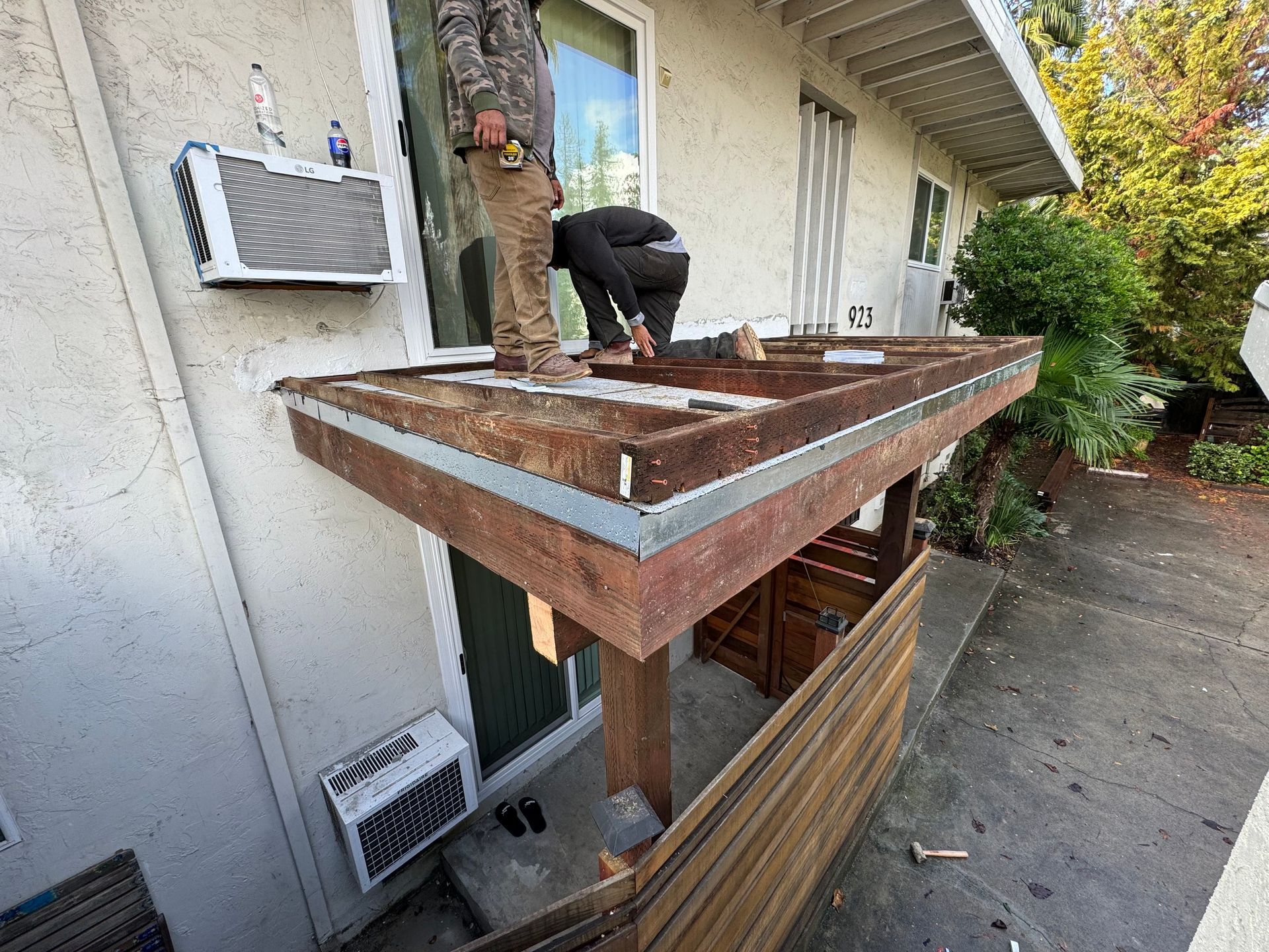 Two people constructing a wooden patio roof attached to a building's exterior.