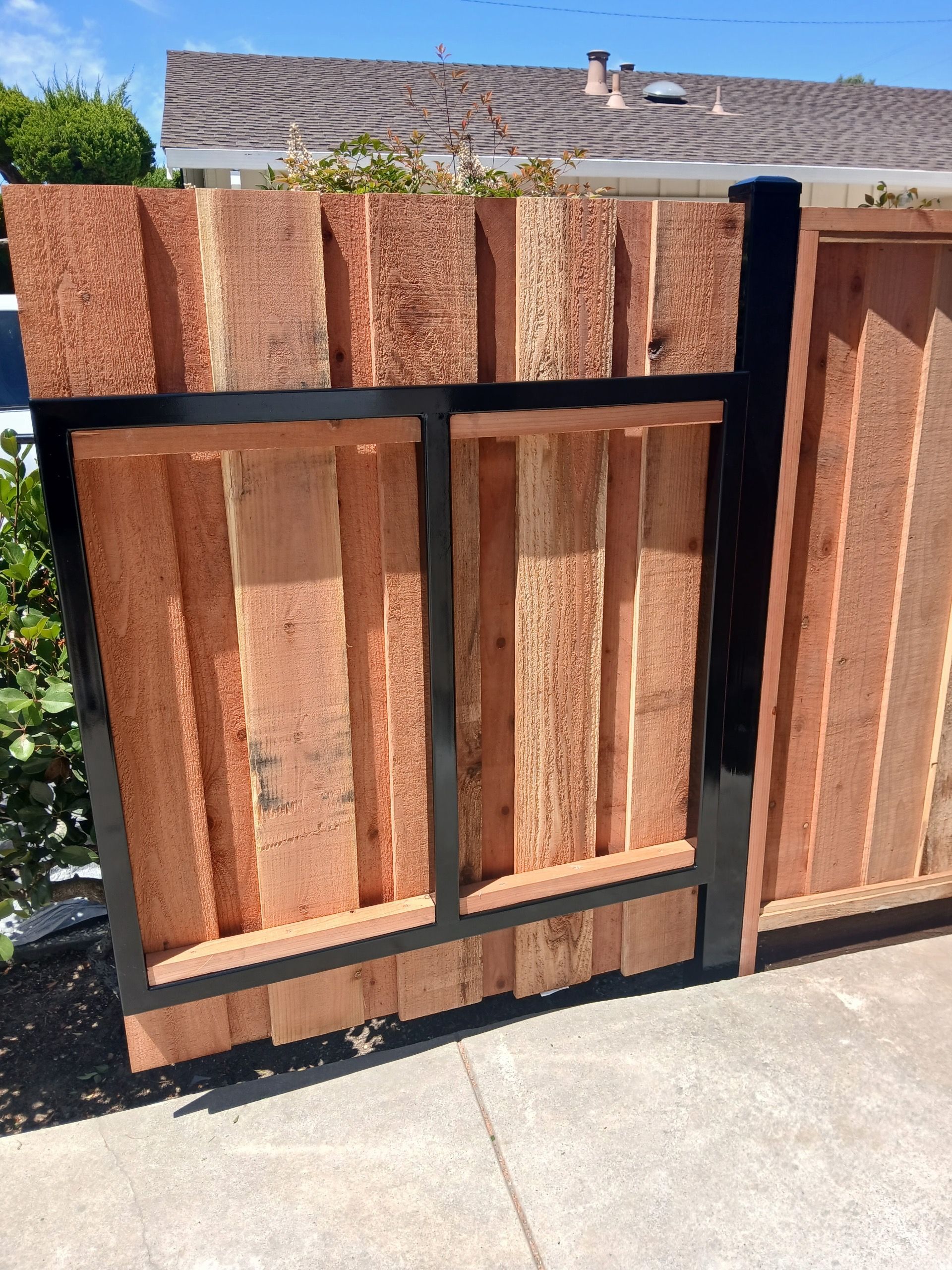 A wood fence section with vertical planks and a black metal support frame, installed on a concrete sidewalk.