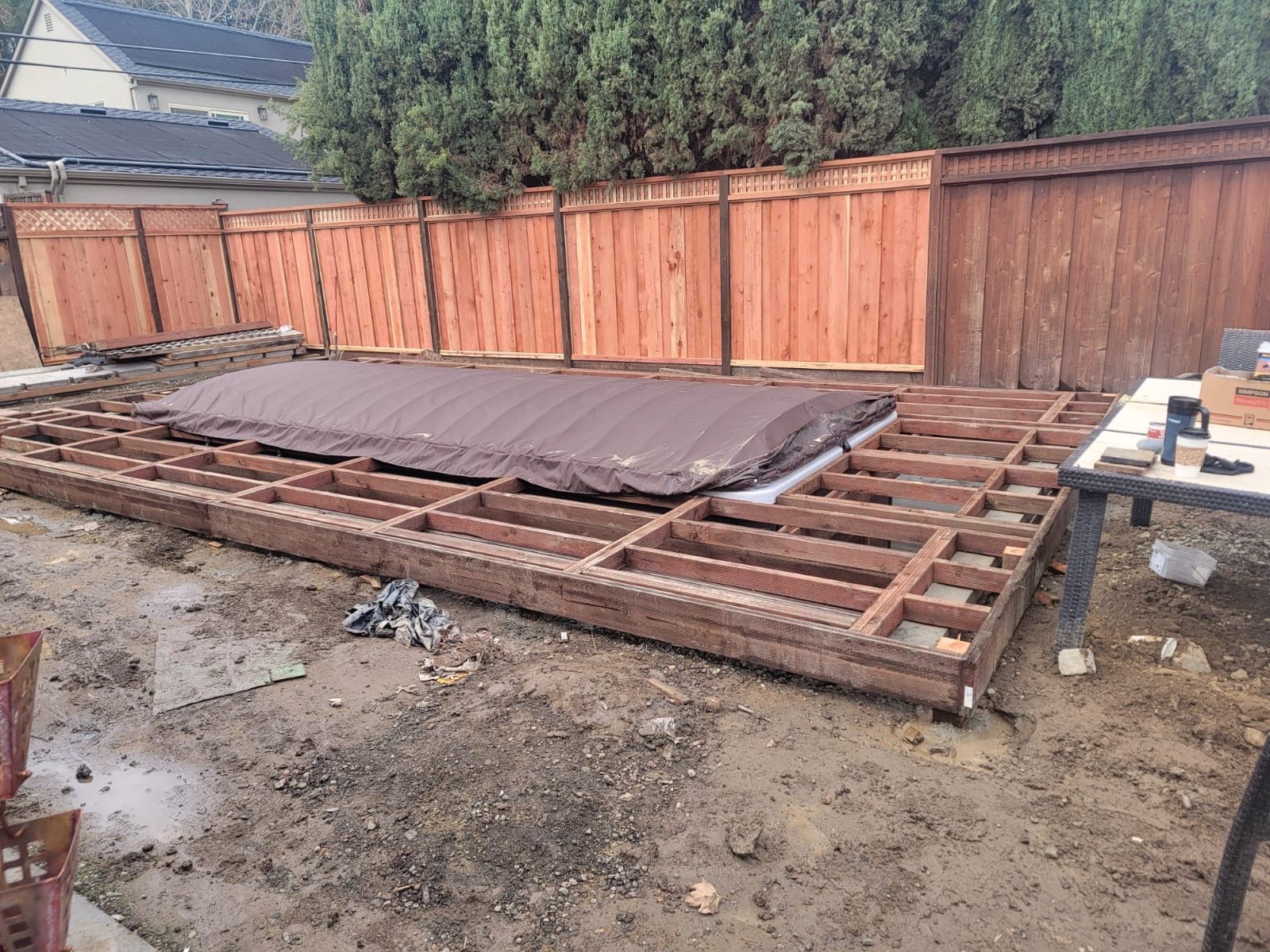 Wooden hot tub deck under construction, covered hot tub, brown fence in backyard.