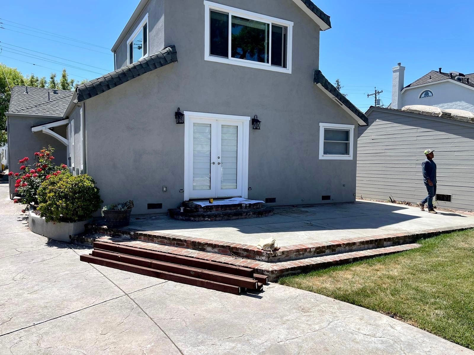 A gray house with white trim, a concrete patio, brick steps, and a person walking in the background on a sunny day.