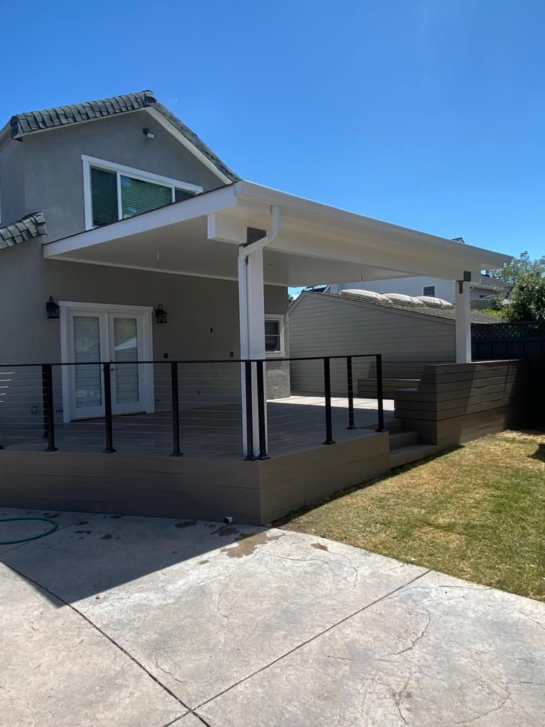 A light gray house with a modern covered patio deck featuring a white roof, black railings, and stone-colored stairs.