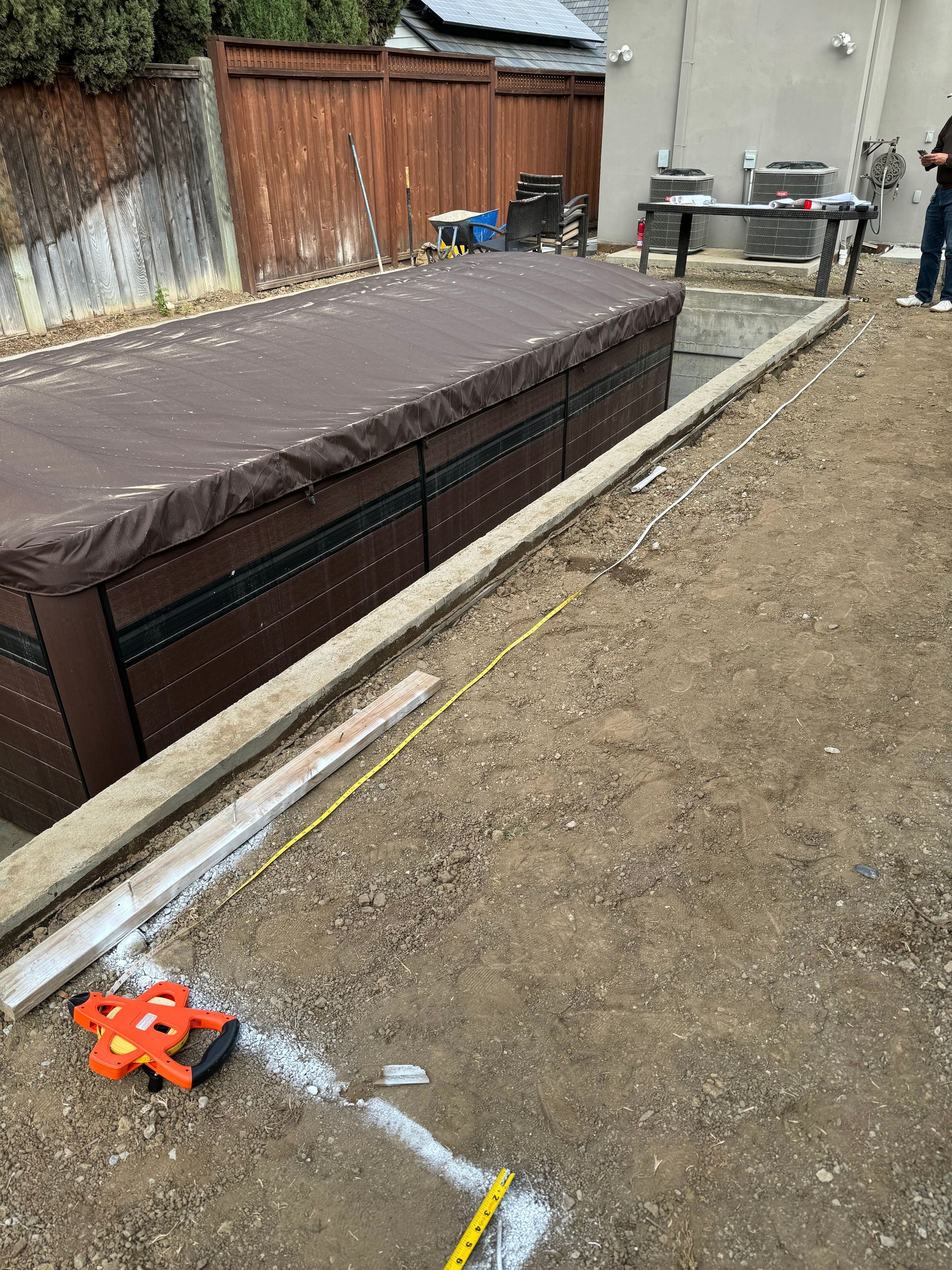 Hot tub covered with brown lid, on gravel surface. Concrete border and construction tools are visible.