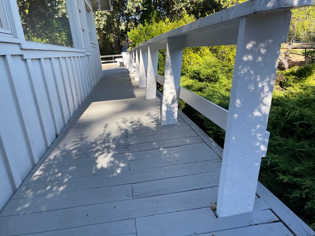 A white wooden porch walkway with a railing, bordered by green bushes on a sunny day.