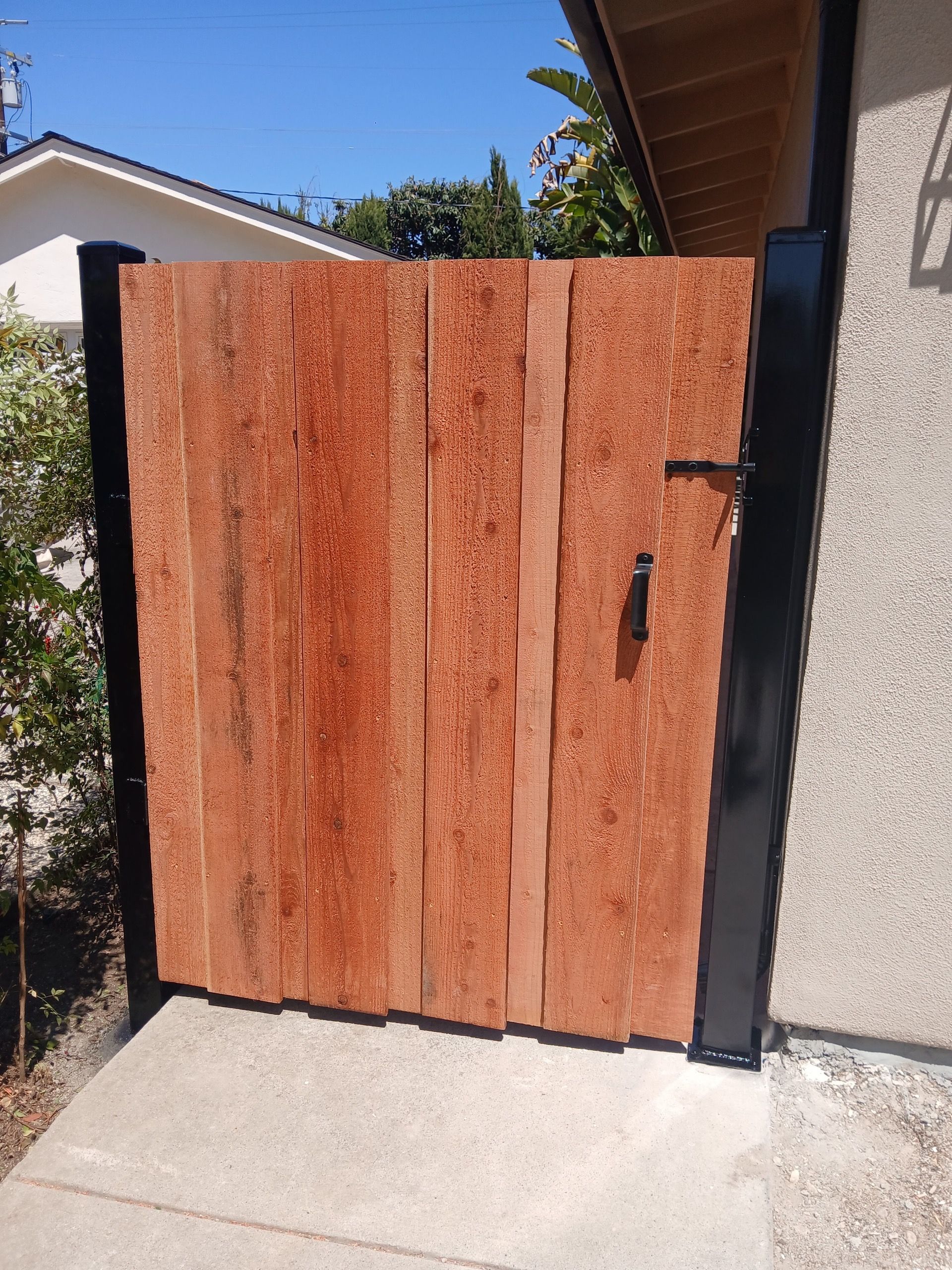 A wooden gate with vertical planks and a black metal frame stands on a concrete path outdoors.
