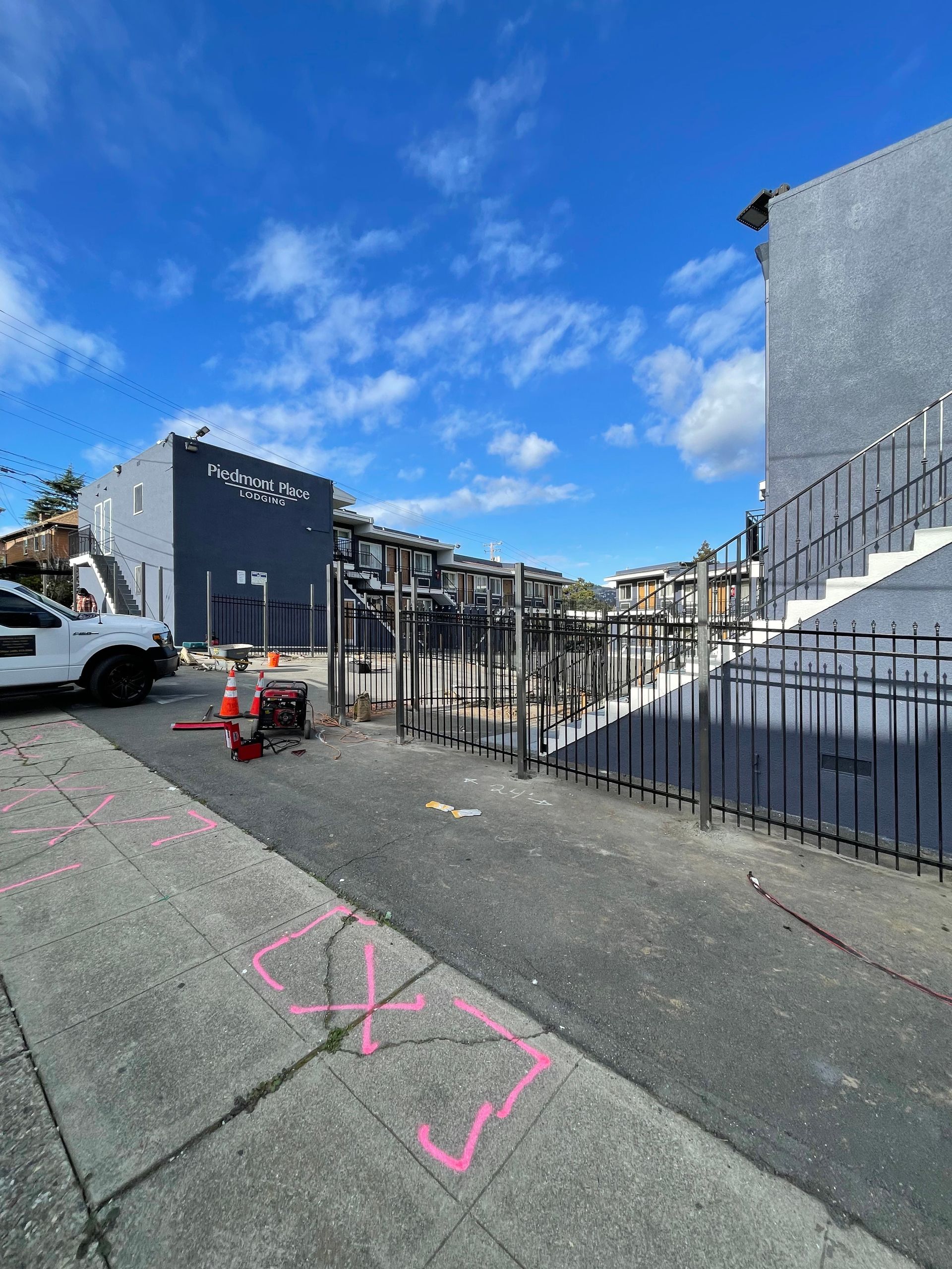 A parking area with gray apartment buildings, a black metal fence, a white pickup truck, and pink markings on the pavement.