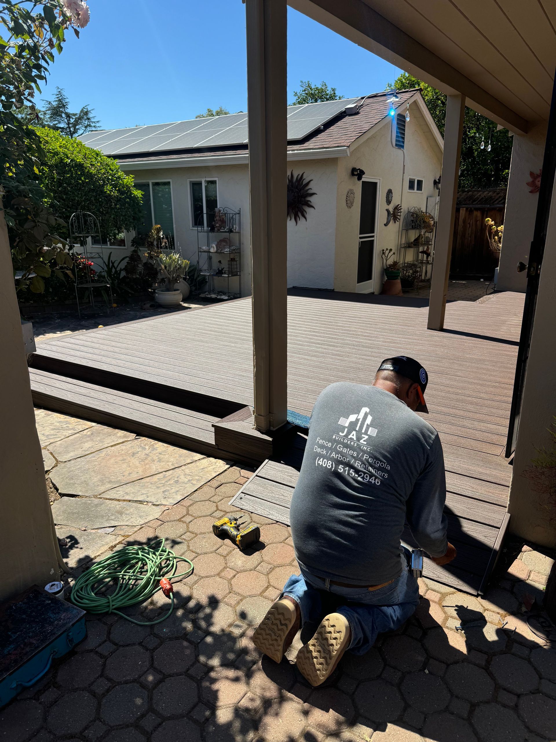 Person kneeling on patio, working on a structure. Patio with circular brick design, house with solar panels in background.