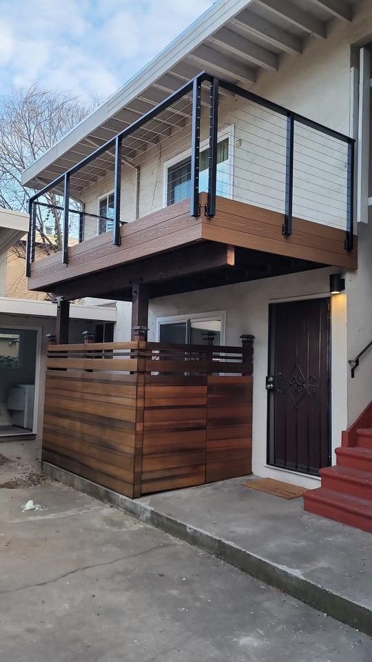 A two-story building exterior with a modern wood-decked balcony and matching privacy screen, beside a brown entry door.