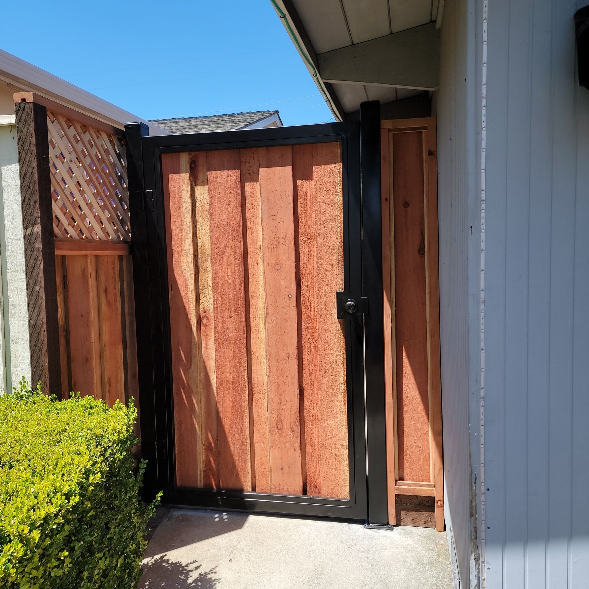 A wooden gate with a black metal frame installed between a house wall and a fence on a concrete walkway.