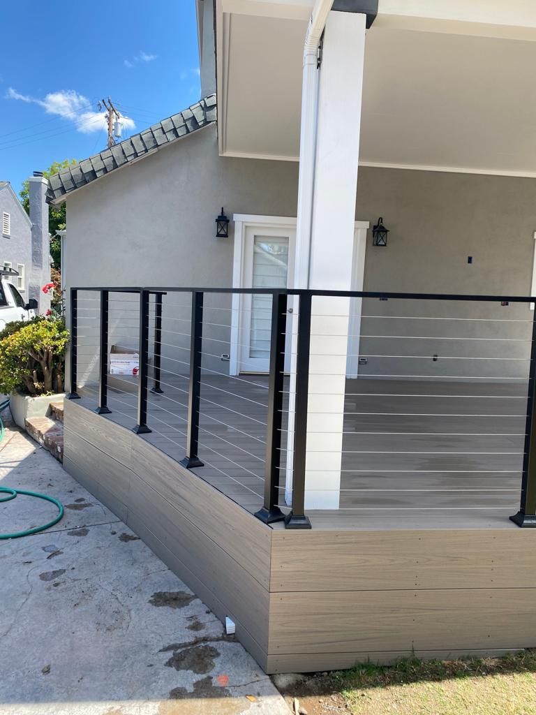 A ground-level view of a newly built deck with dark railings and horizontal cables against a gray house exterior.