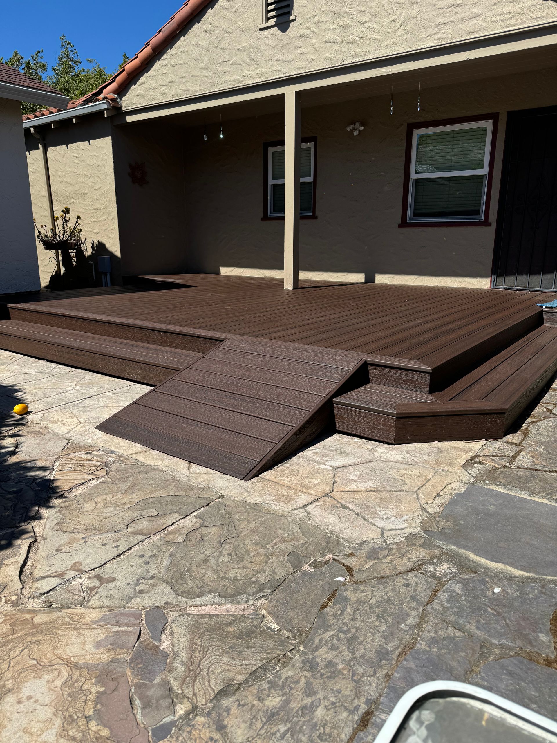 A brown composite deck with stairs and a ramp leads to a house with a stone patio.