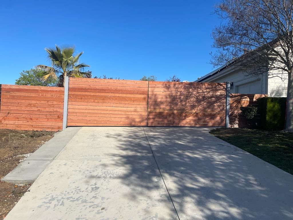 A wide, horizontal wooden slat gate spans a concrete driveway in front of a house under a clear blue sky.