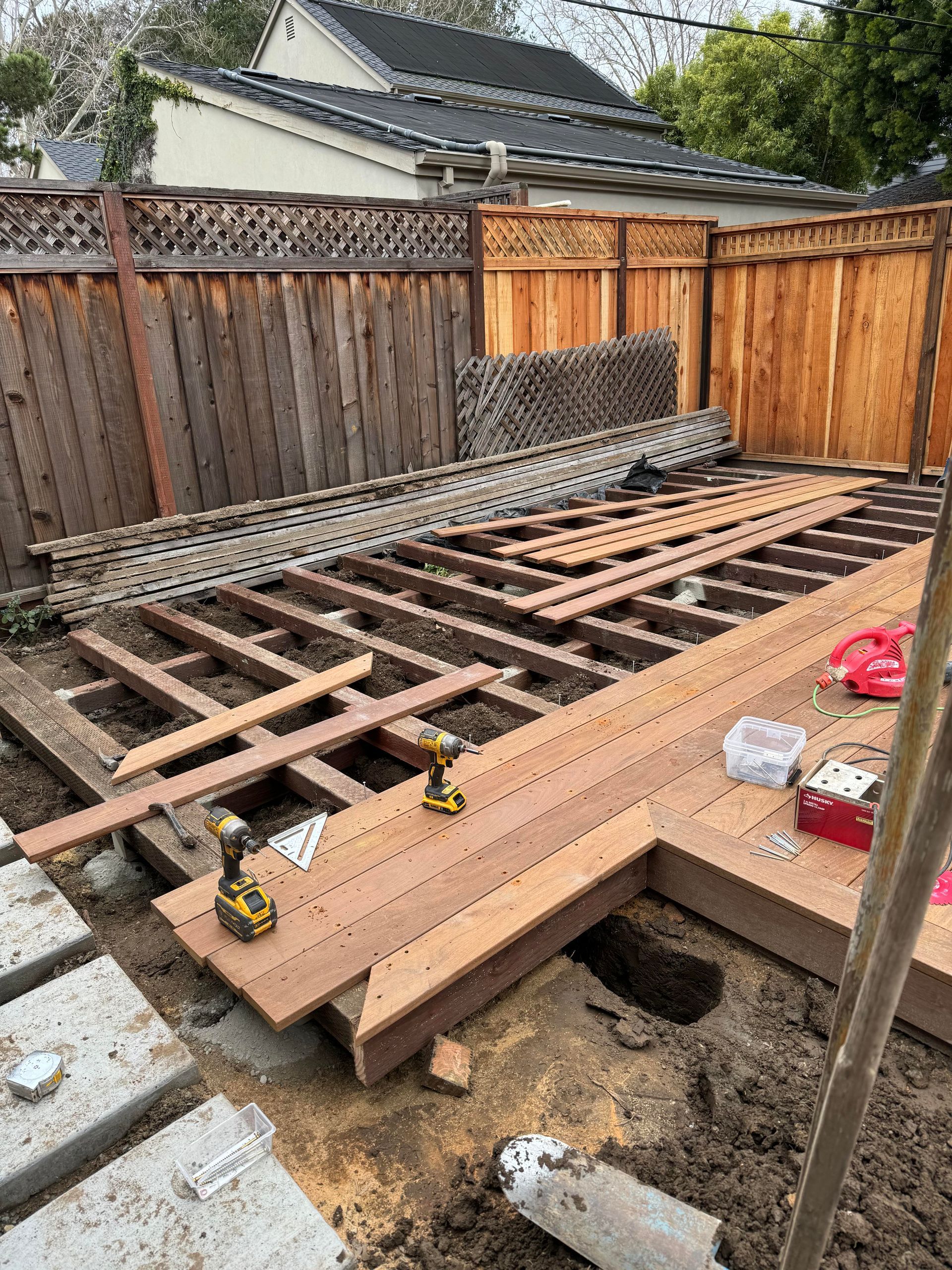 Construction of a wooden deck in a backyard. Brown wood, tools, and dirt are visible. Fence in the background.