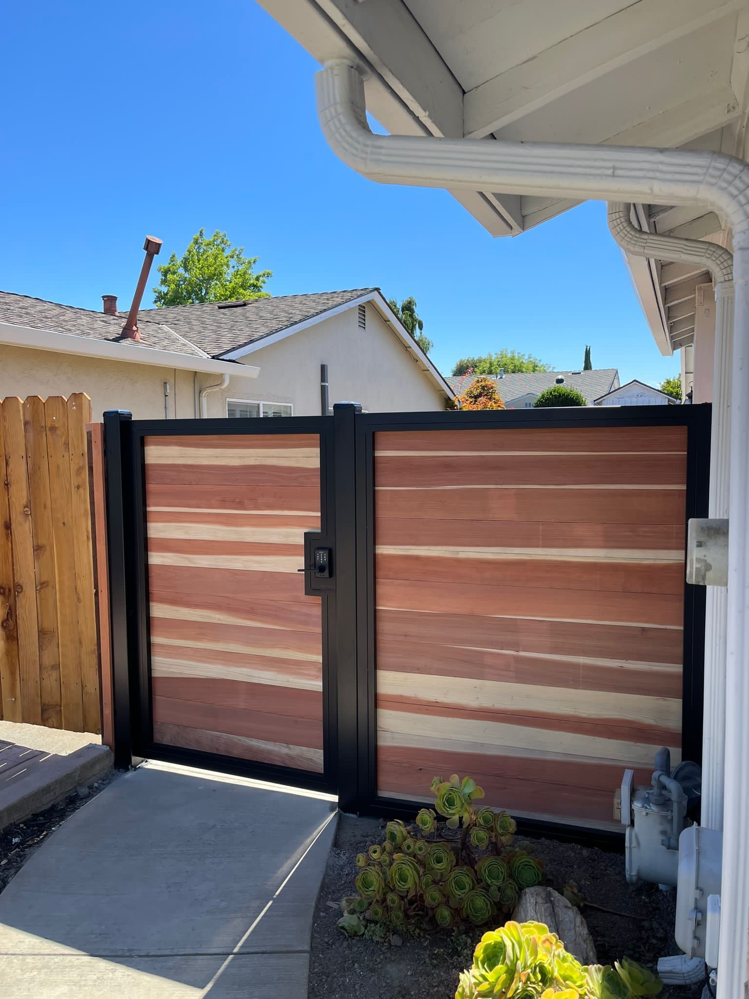 A modern gate featuring horizontal wood panels framed in black metal, installed next to a house under a clear blue sky.