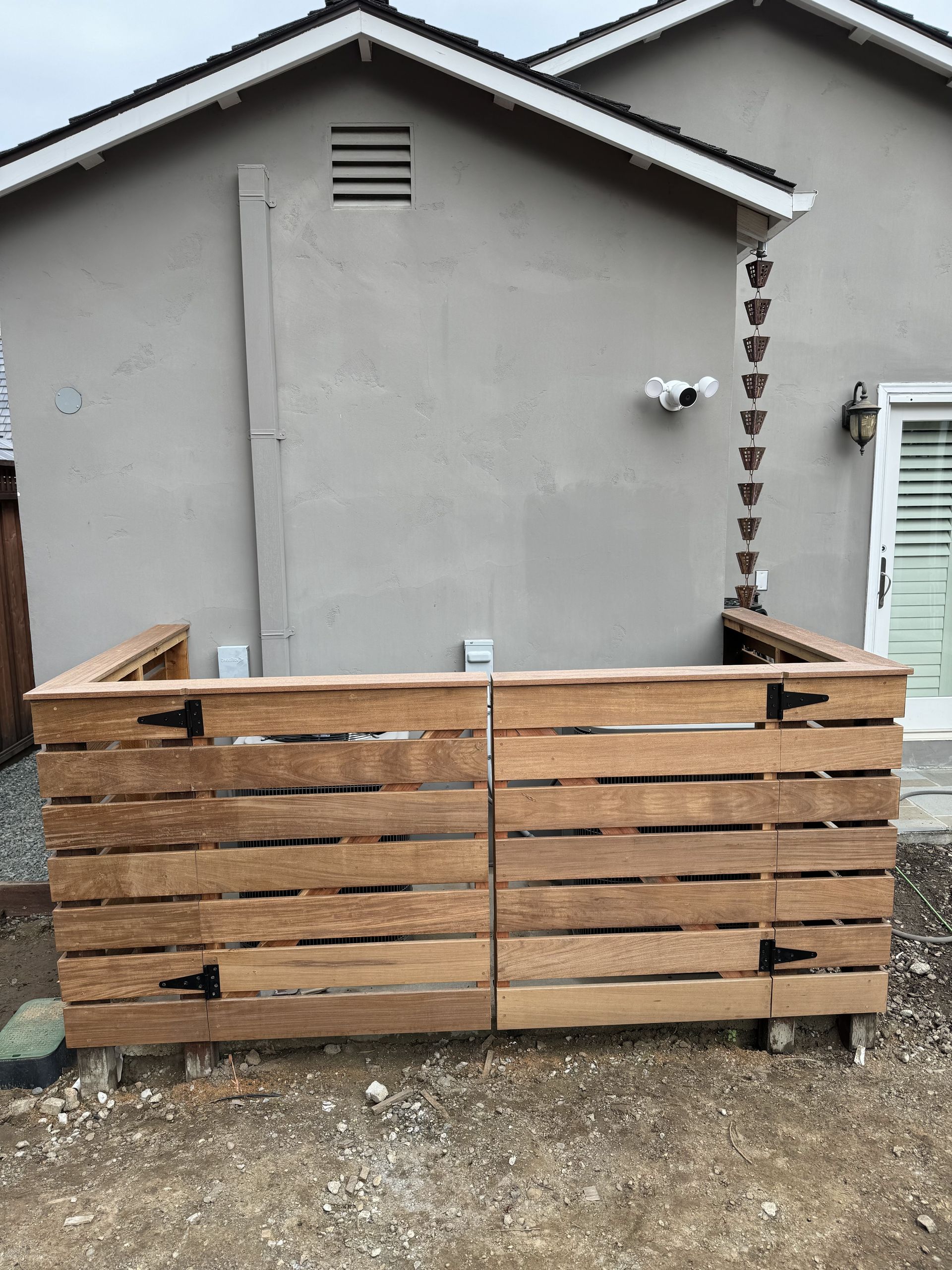 Wooden bin enclosure against a house, two doors, black hinges, a security camera, and a rain chain.