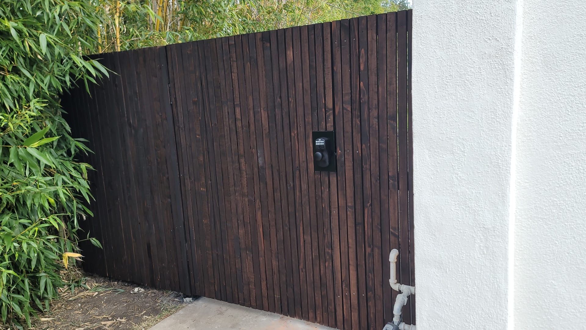 A dark wooden slatted gate stands next to a white stucco wall, framed by green bamboo foliage.