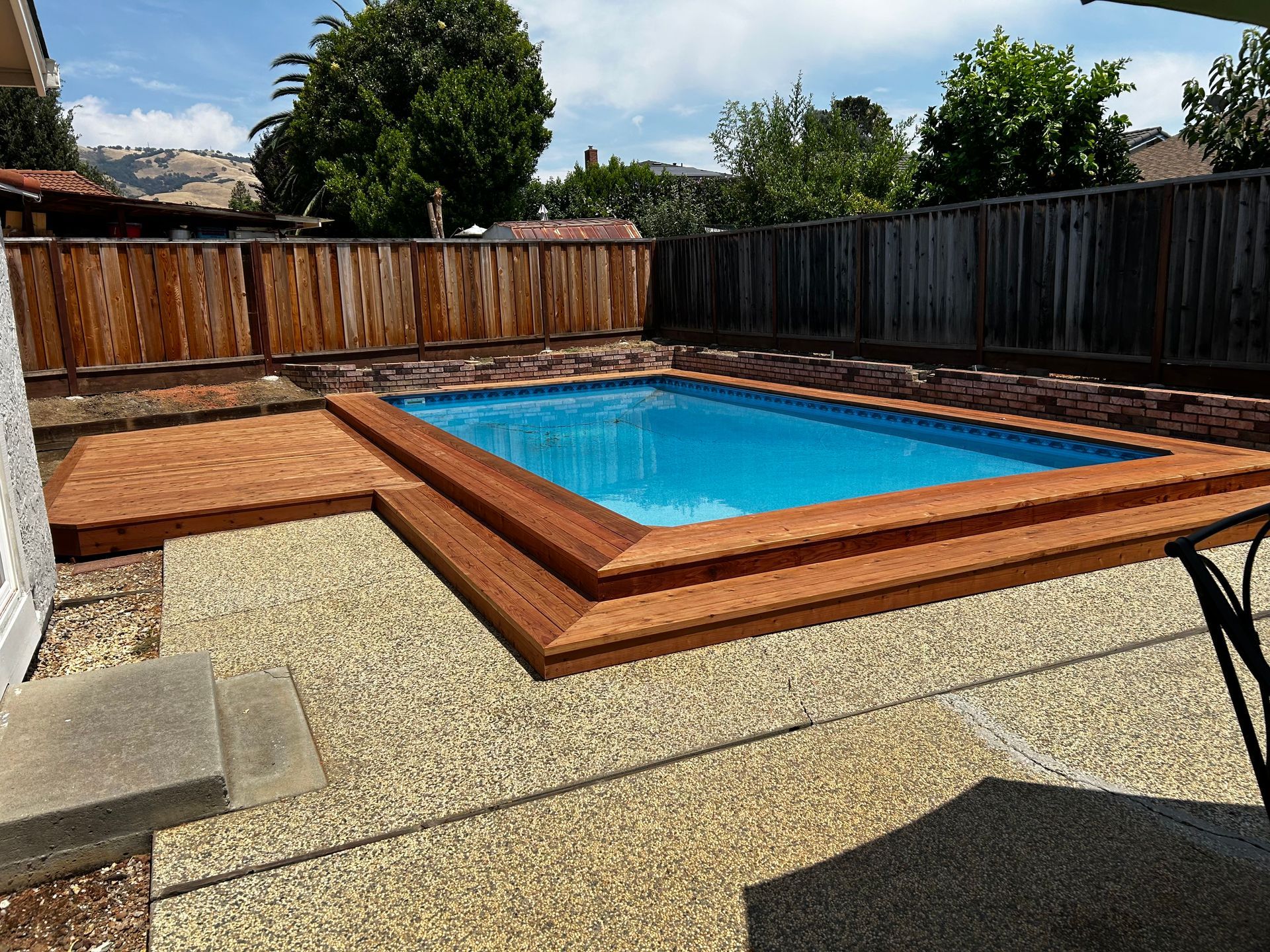 Small rectangular pool with wooden steps, surrounded by a pebble patio and wooden fence.