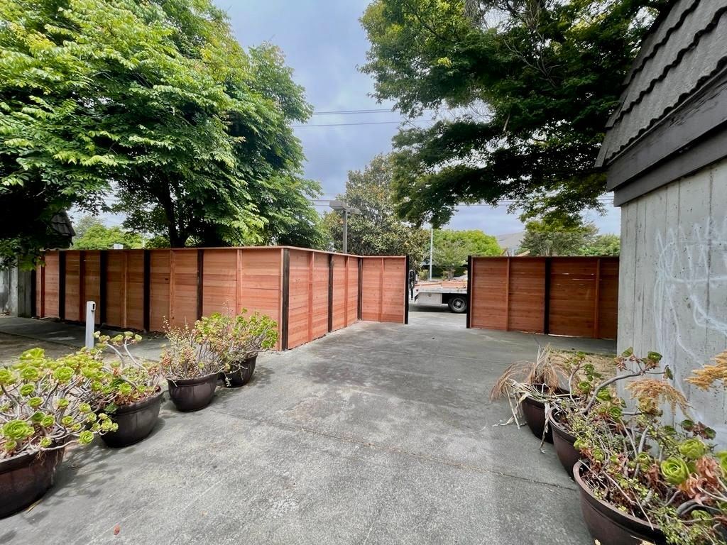 Wooden fence with open gate, concrete path, and potted plants. Trees and overcast sky in background.