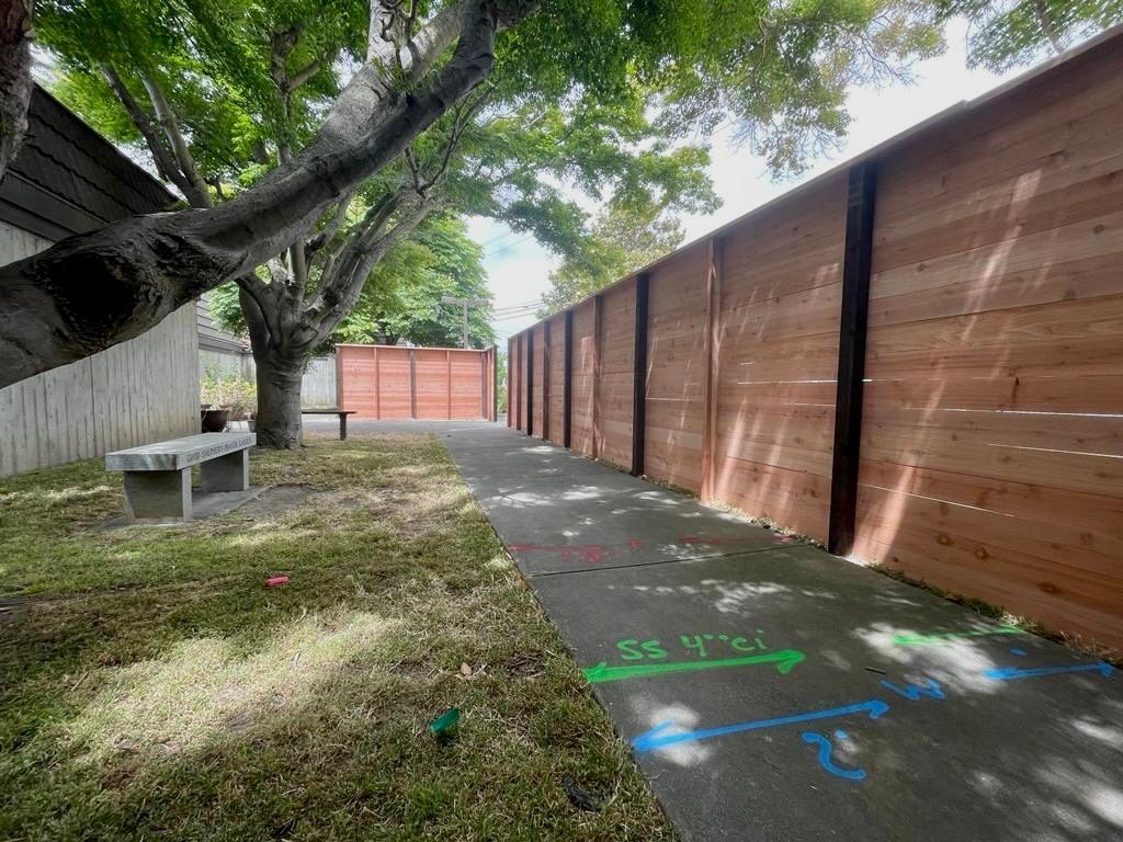A sidewalk runs beside a wooden fence and a grassy area with a bench under a large tree, marked with colorful ground arrows.