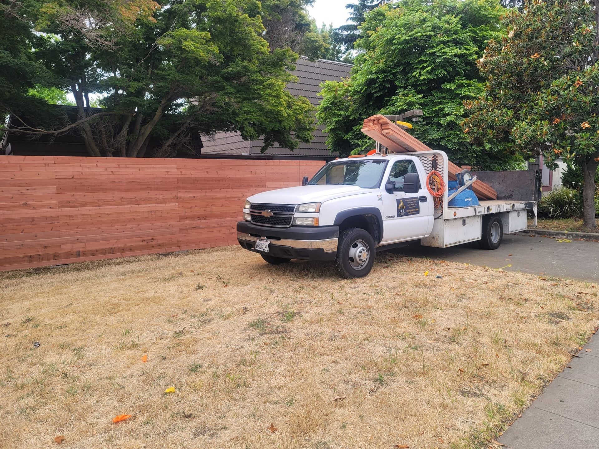 A white service truck parked on dry grass in front of a new wooden fence and lush green trees.