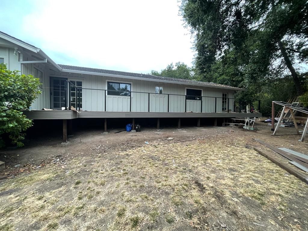 Exterior view of a light-colored house with a raised deck, against a backdrop of trees and a dry, grassy yard.