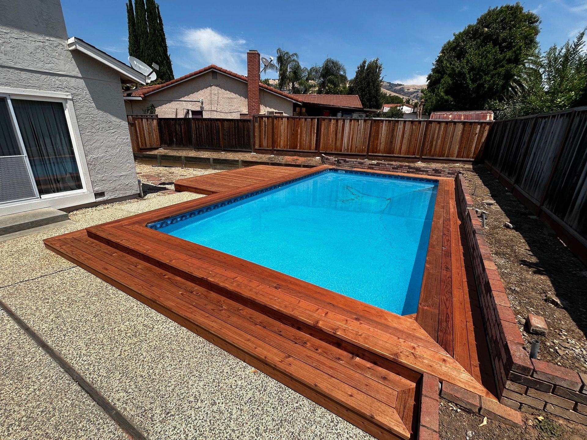 Rectangular in-ground pool surrounded by a wooden deck and brick, nestled between a house and wooden fence.