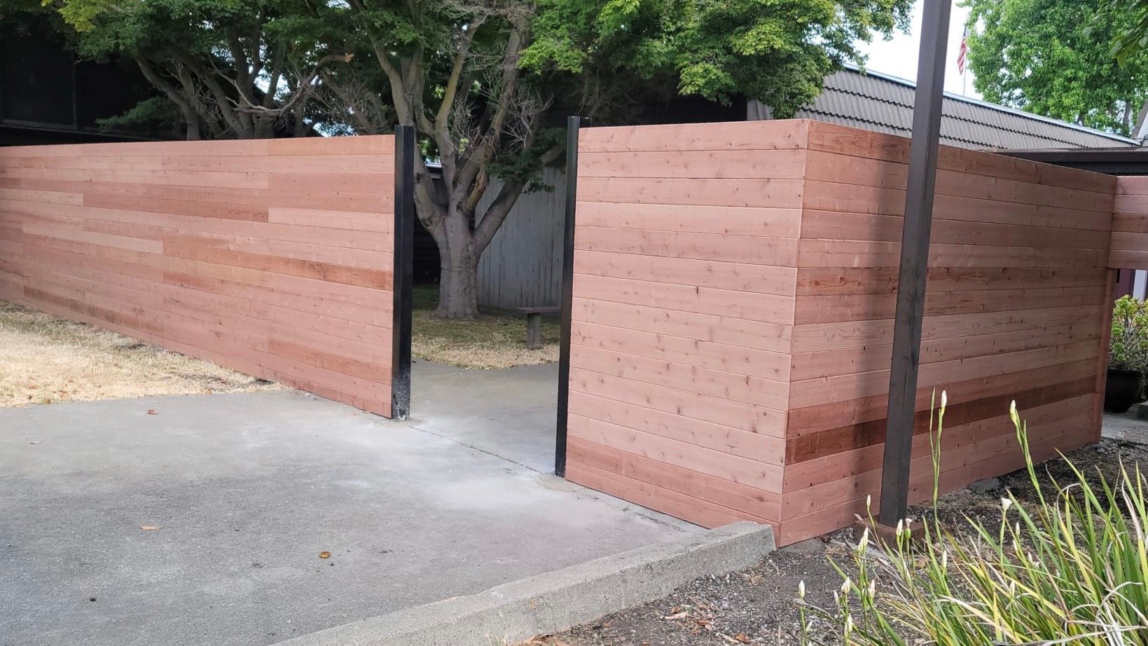A horizontal wooden slat privacy fence with a central gate opening over a concrete driveway, surrounded by green trees.