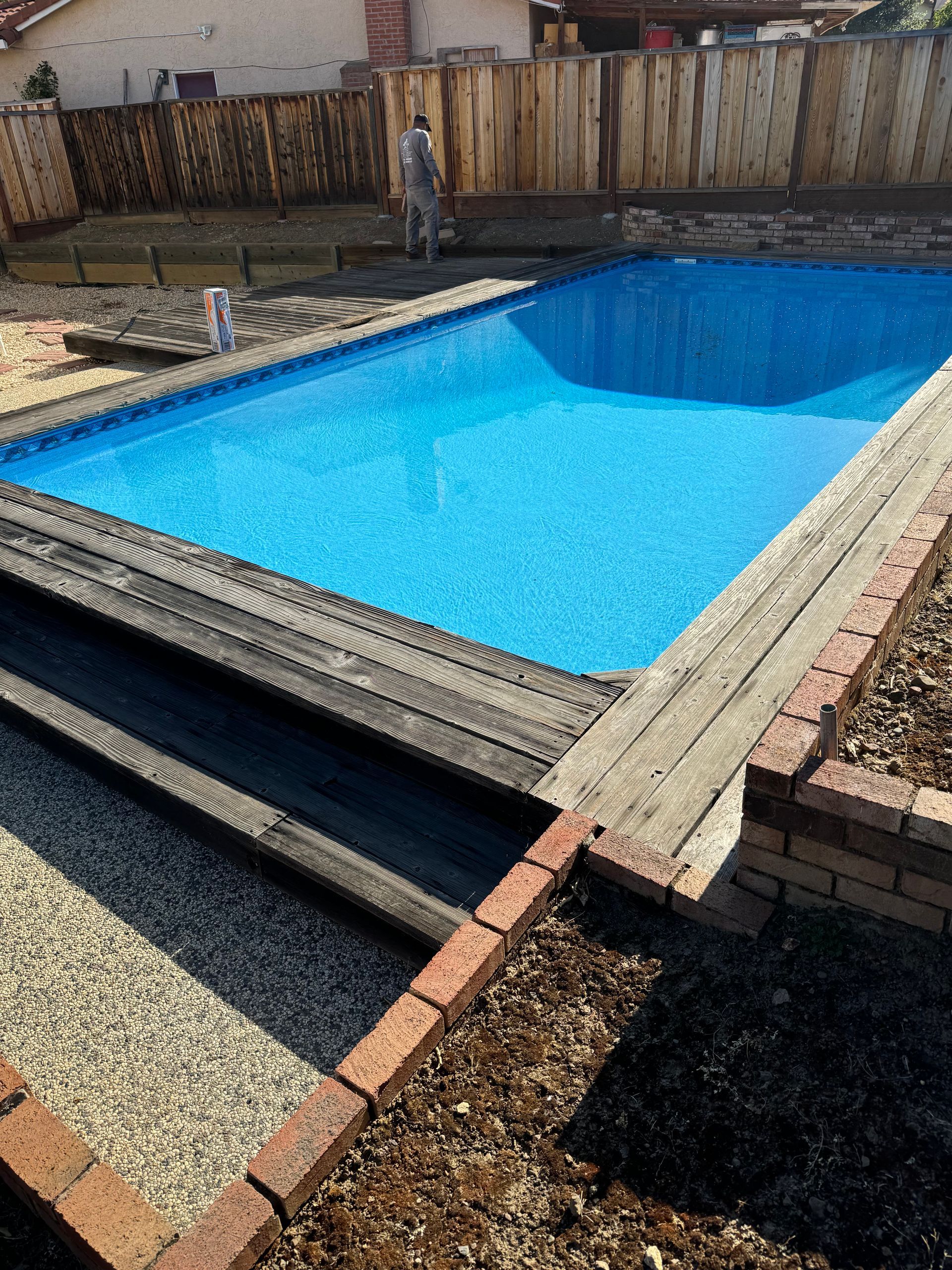 Rectangular blue pool surrounded by a wooden deck and brick edging. A person is standing in the background.