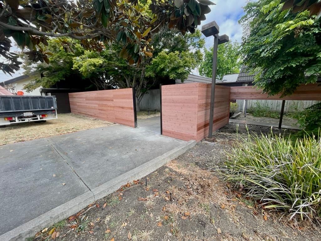 A modern wood-slat fence with an open entry gate on a concrete driveway, surrounded by trees and landscaping.