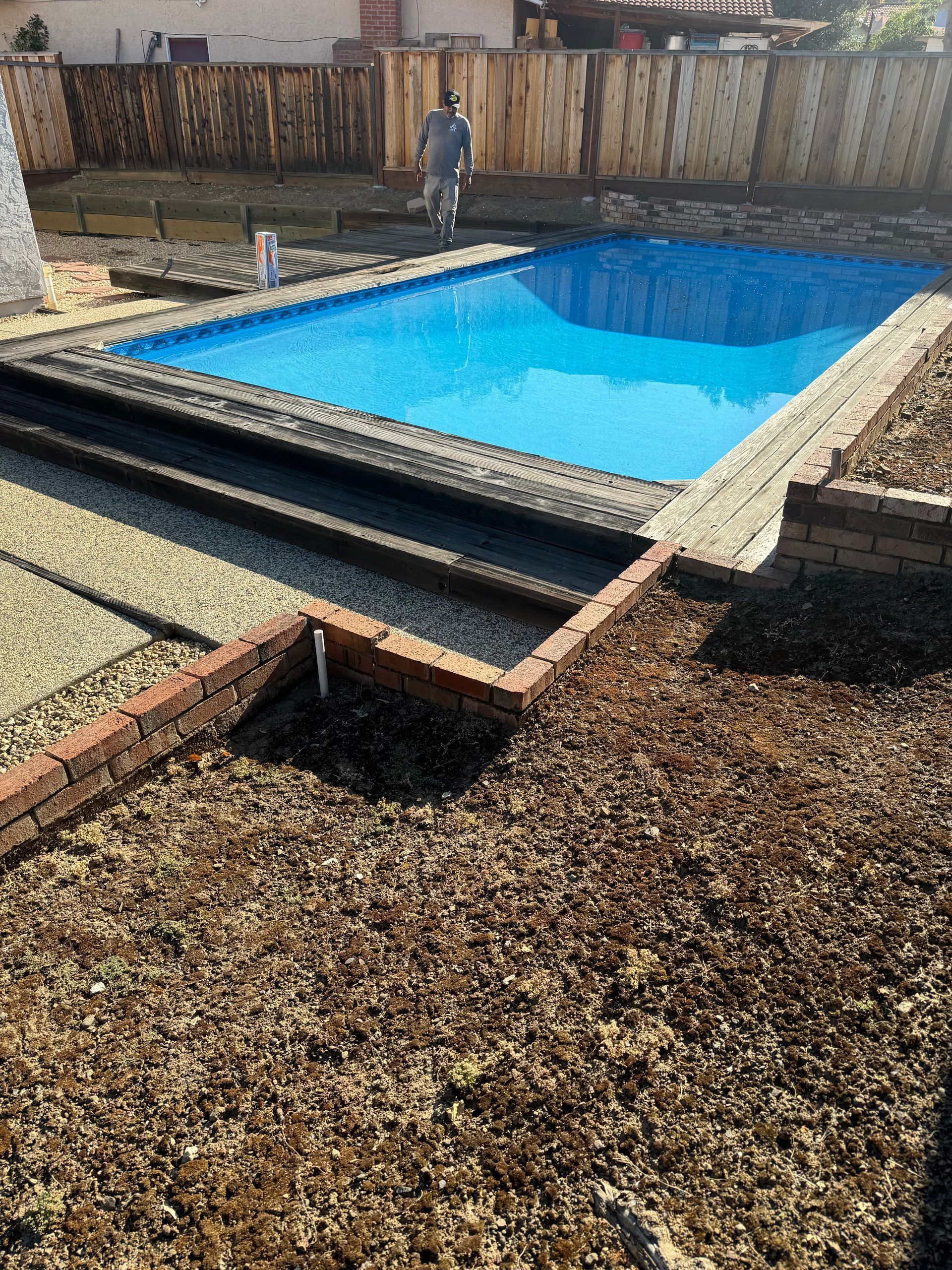 Backyard pool with blue water, brick edging, and a wooden fence. A person walks near the pool.