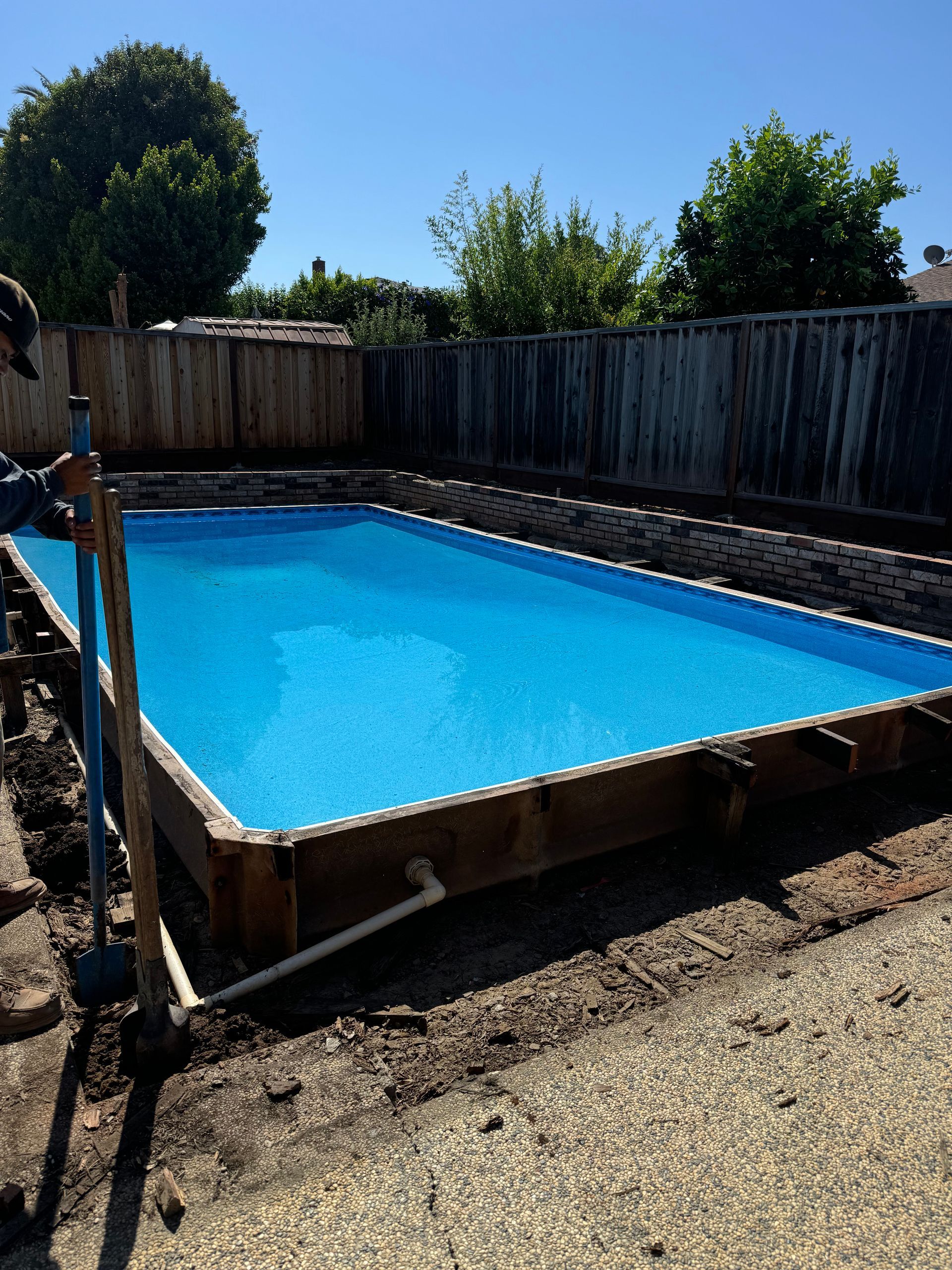 Above-ground rectangular pool with bright blue water, surrounded by wooden framing and gravel, in a backyard.
