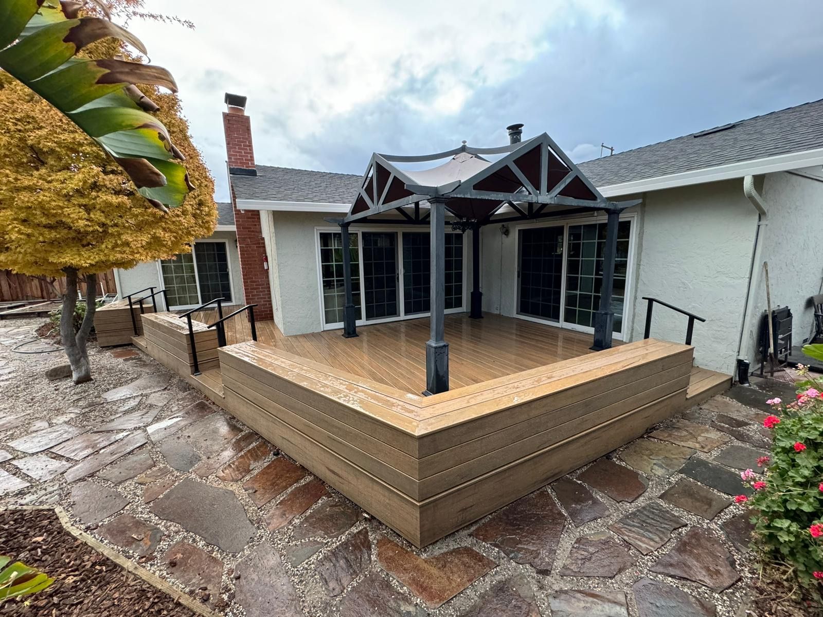 A tan composite deck with a metal gazebo, set against a house with a red brick chimney and stone patio.
