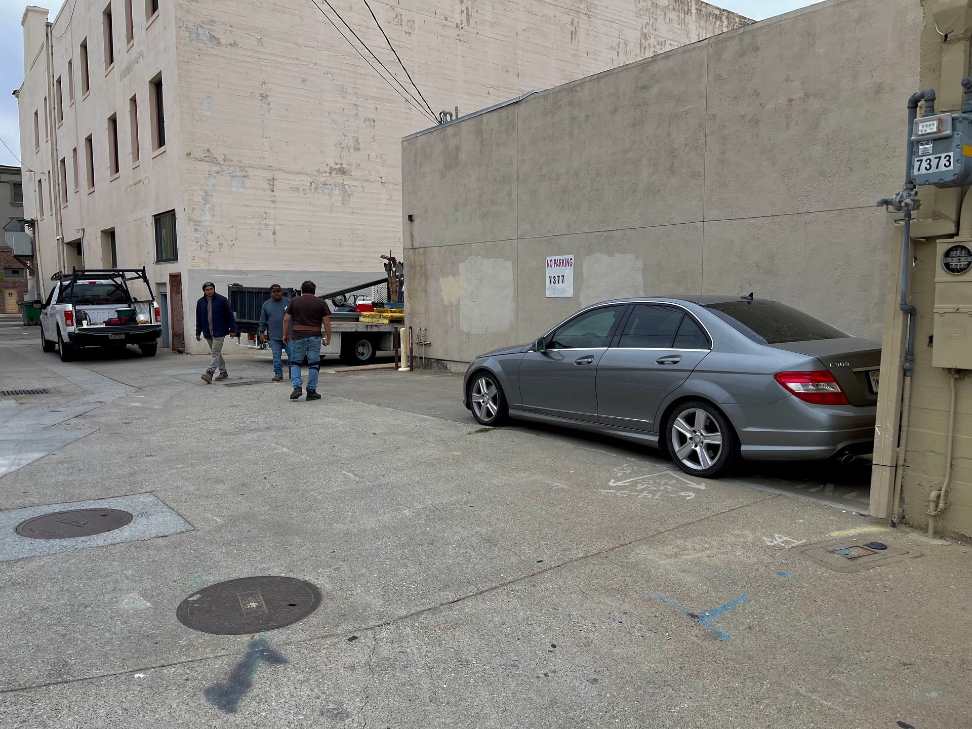 Alley scene: Gray car parked near wall. Several people by truck. Building in background. Cloudy sky.
