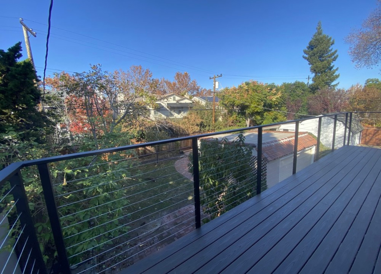 Deck with black railing overlooking a yard with trees and houses, under a blue sky.