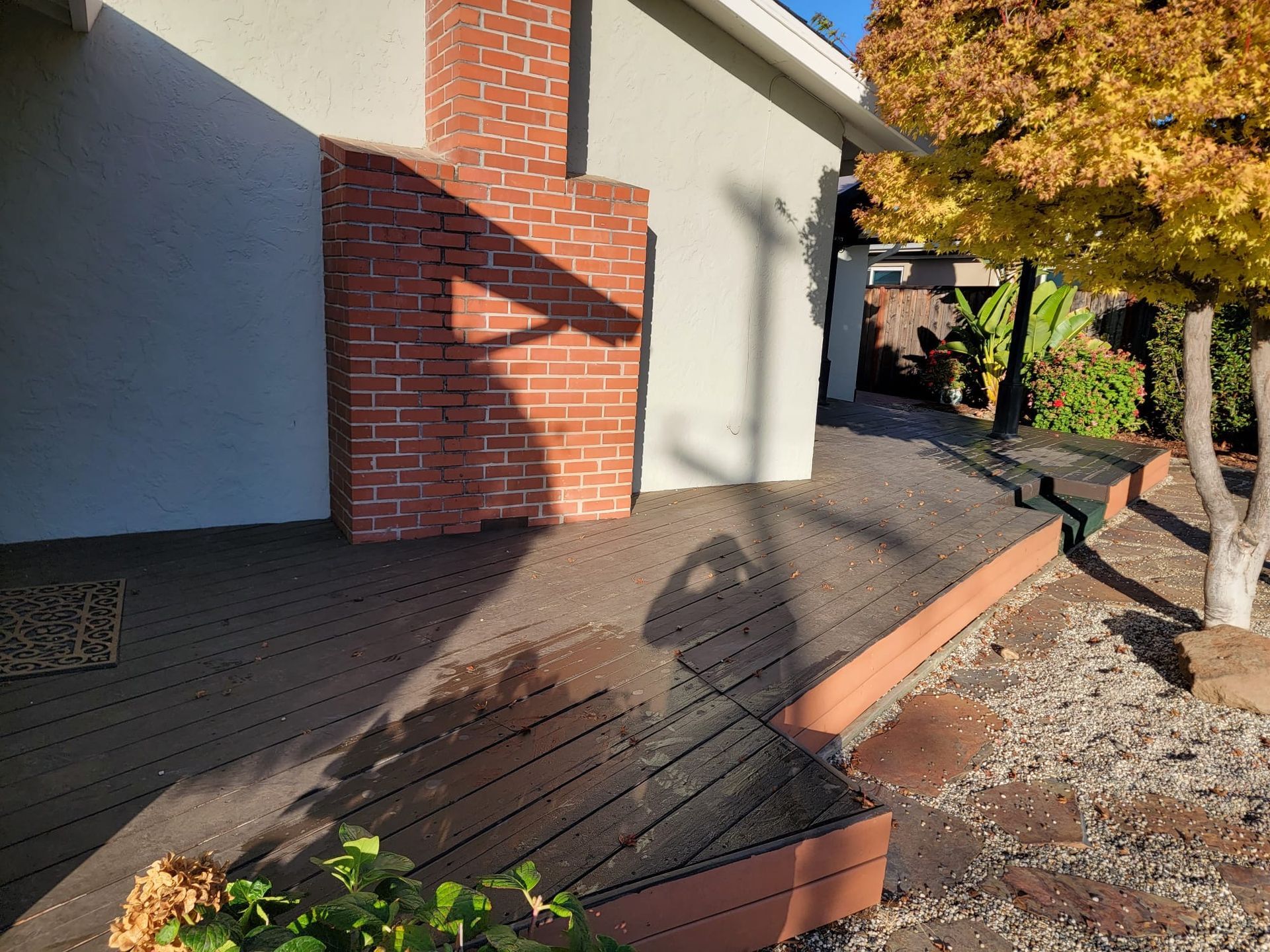 A brown wood deck wraps around a brick chimney on a house exterior, with a yellow tree and gravel yard nearby.
