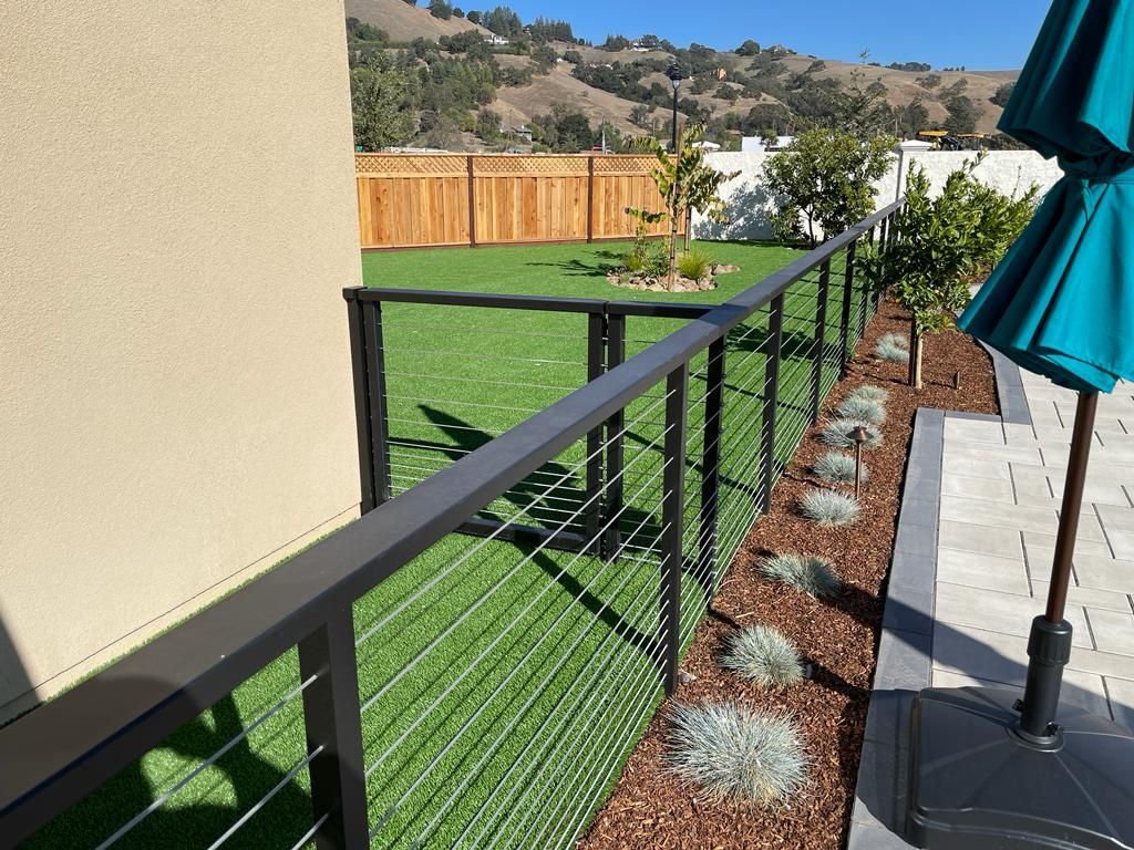 Black cable railing next to a garden bed with gray plants. Green lawn, wooden fence, and hills in background.