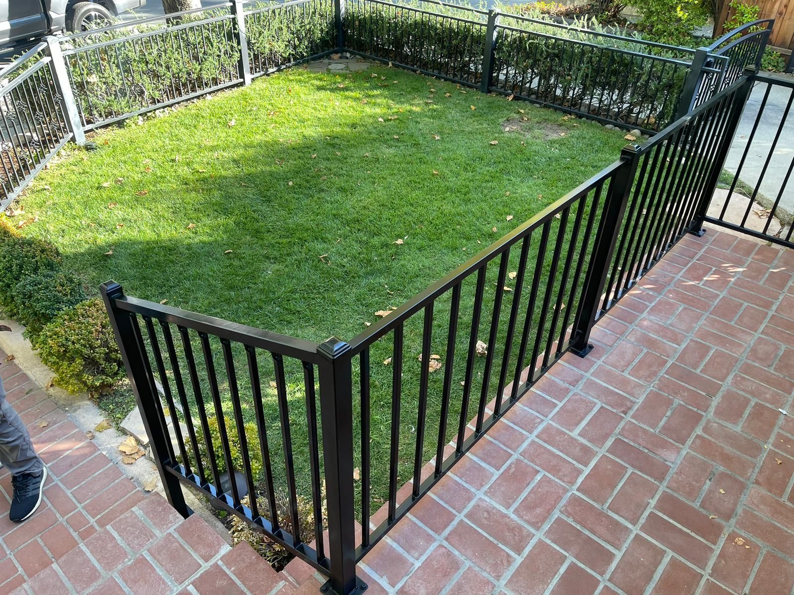 Black fenced small yard with green grass, surrounded by brick patio and hedges.