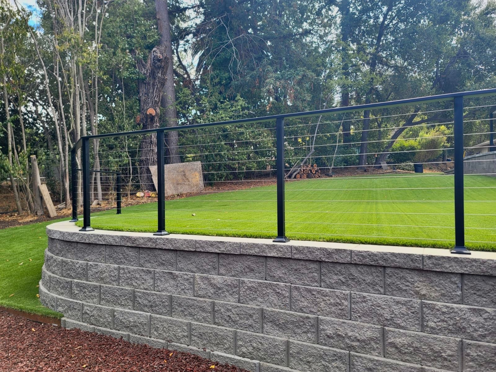 Black metal fence atop a gray stone retaining wall, bordering a grassy yard with trees in the background.