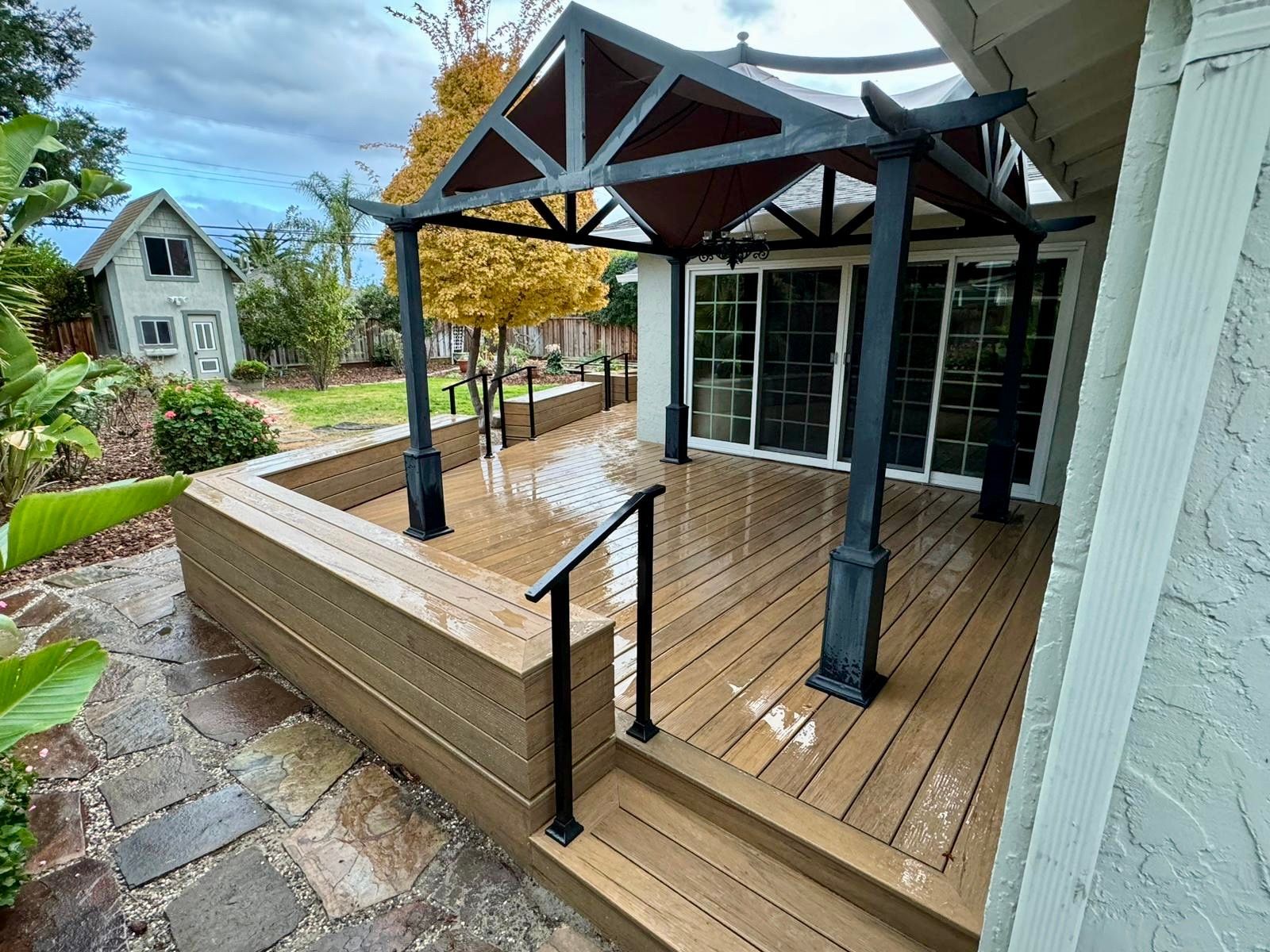 A tan composite deck with a black pergola, a tiered bench, and steps leading to a house, set in a backyard landscape.
