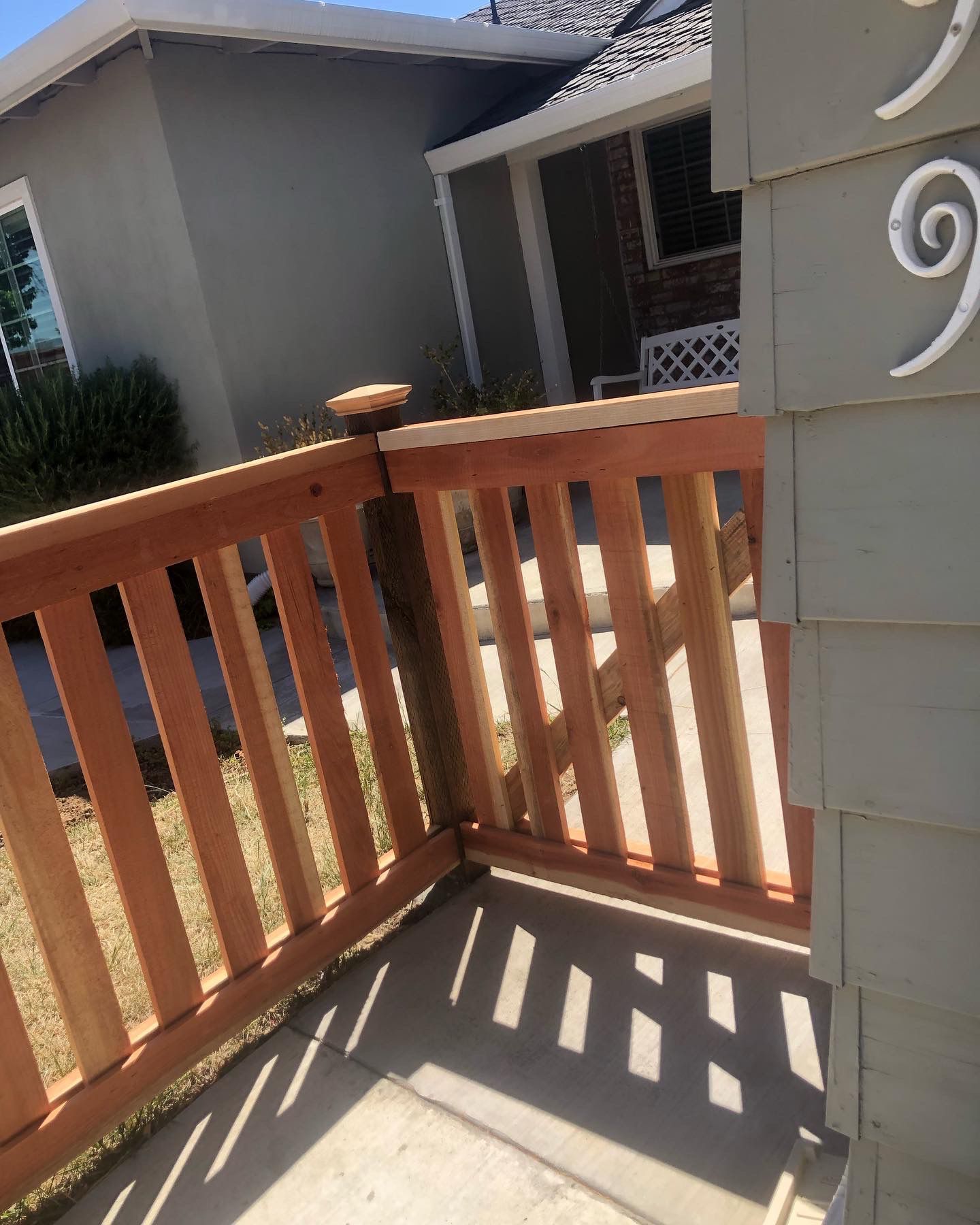 Wooden gate with vertical slats, attached to a gray house, casting shadows on the walkway.