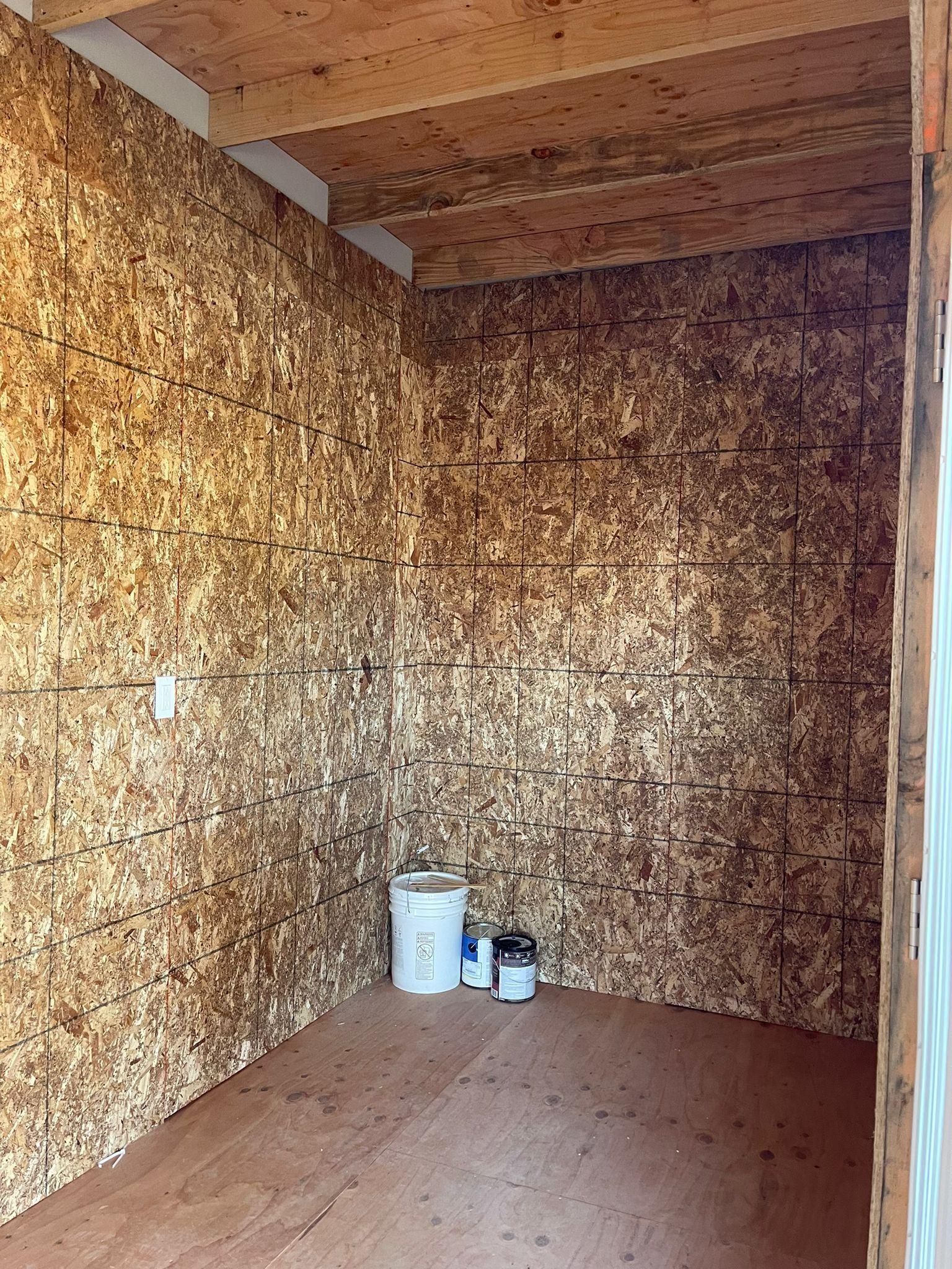 Small interior room with walls and ceiling of brown wood panels; water heater, paint, and other items sit on the floor.